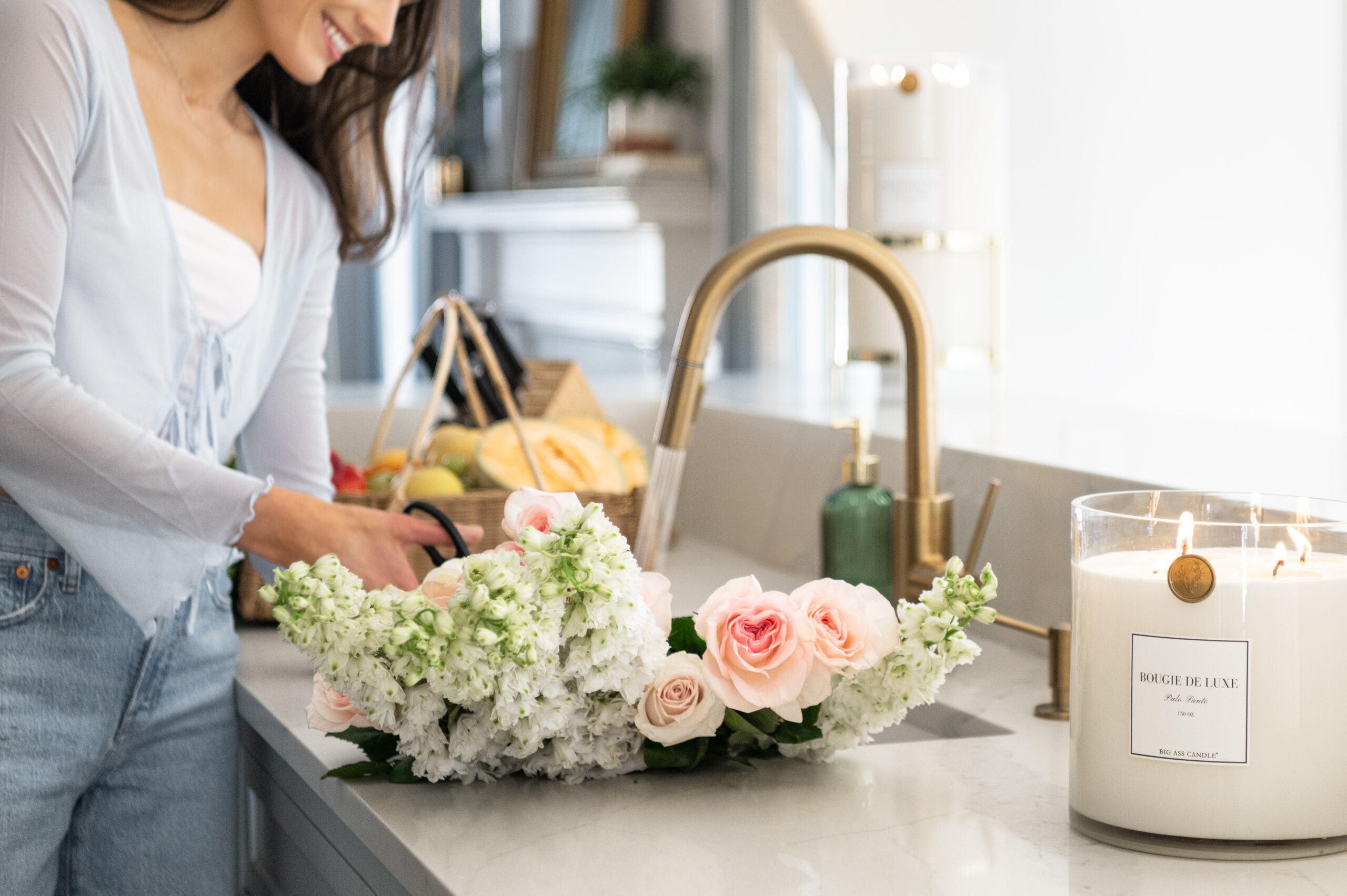 A smiling woman preparing flowers in a kitchen with a luxurious candle lit beside her, capturing the essence of seasonal photography in a relaxed setting.