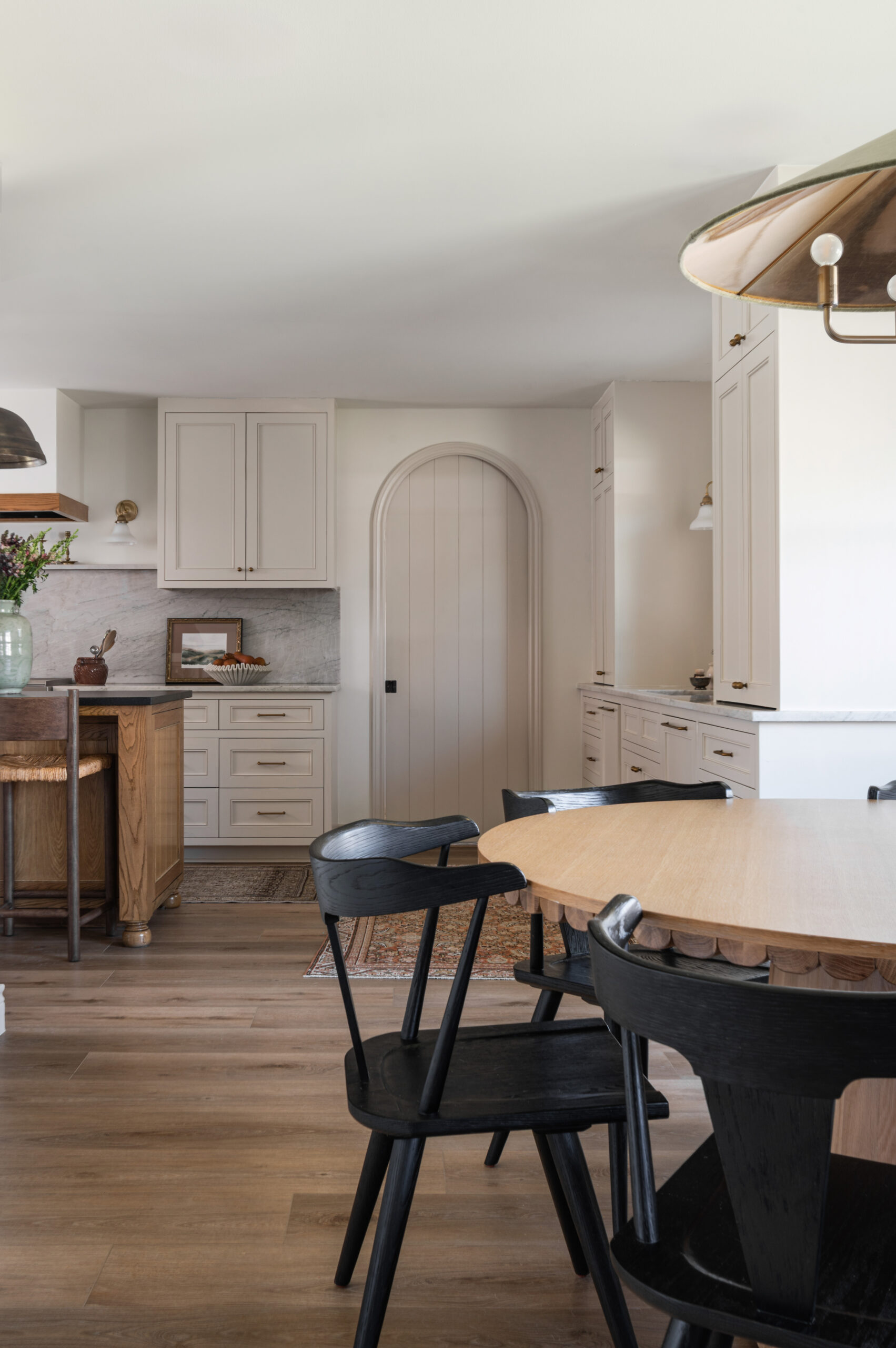 A warm, natural-toned kitchen with creamy cabinetry, a scalloped-edge wood table, and contrasting black chairs, styled with earthy accents and soft lighting.