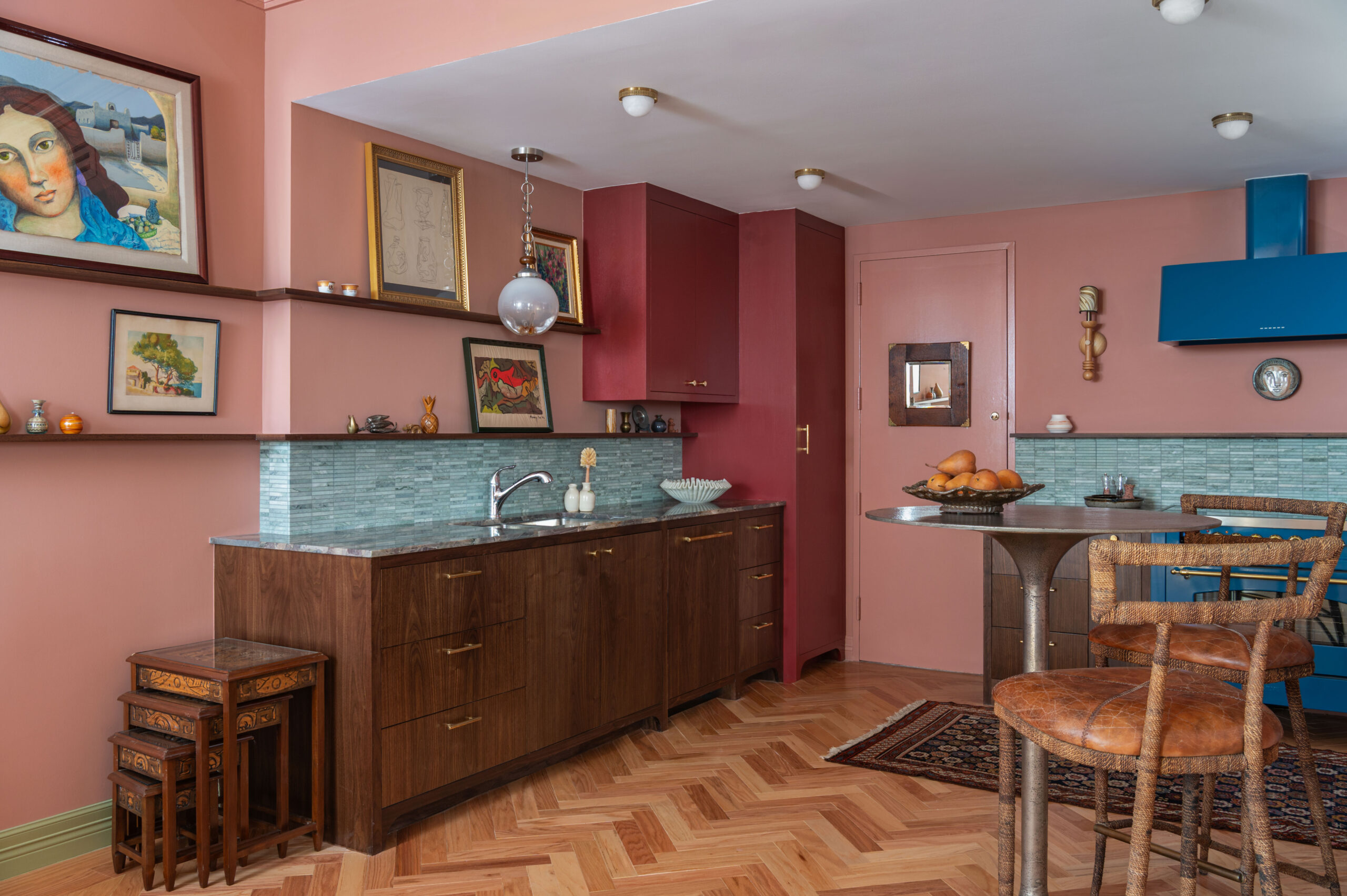 A bold and eclectic kitchen with terracotta pink walls, red cabinetry, and green glass backsplash, enhanced by collected artwork and herringbone floors.