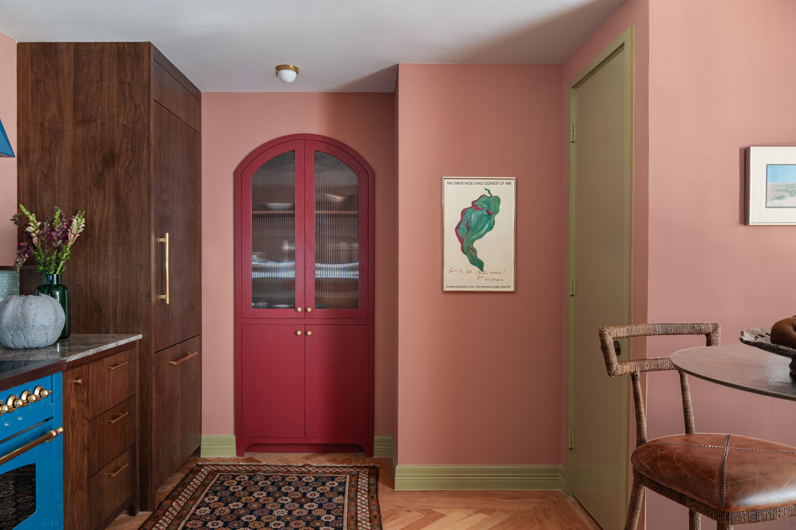 An arched red glass cabinet and olive green trim pop against a pink kitchen wall, a playful combination that highlights how to photograph interior spaces that embrace bold color palettes.