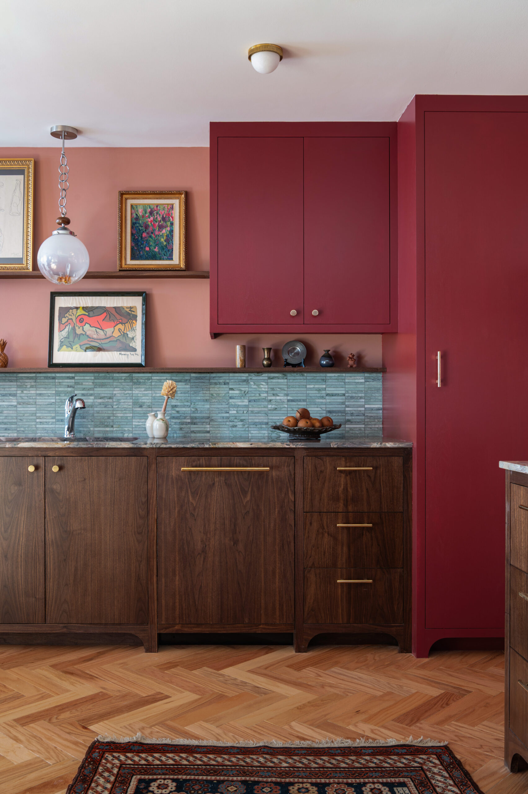 Close-up of a rich kitchen vignette featuring wood cabinetry, brass hardware, red upper cabinets, and a layered gallery shelf with artwork and ceramics.