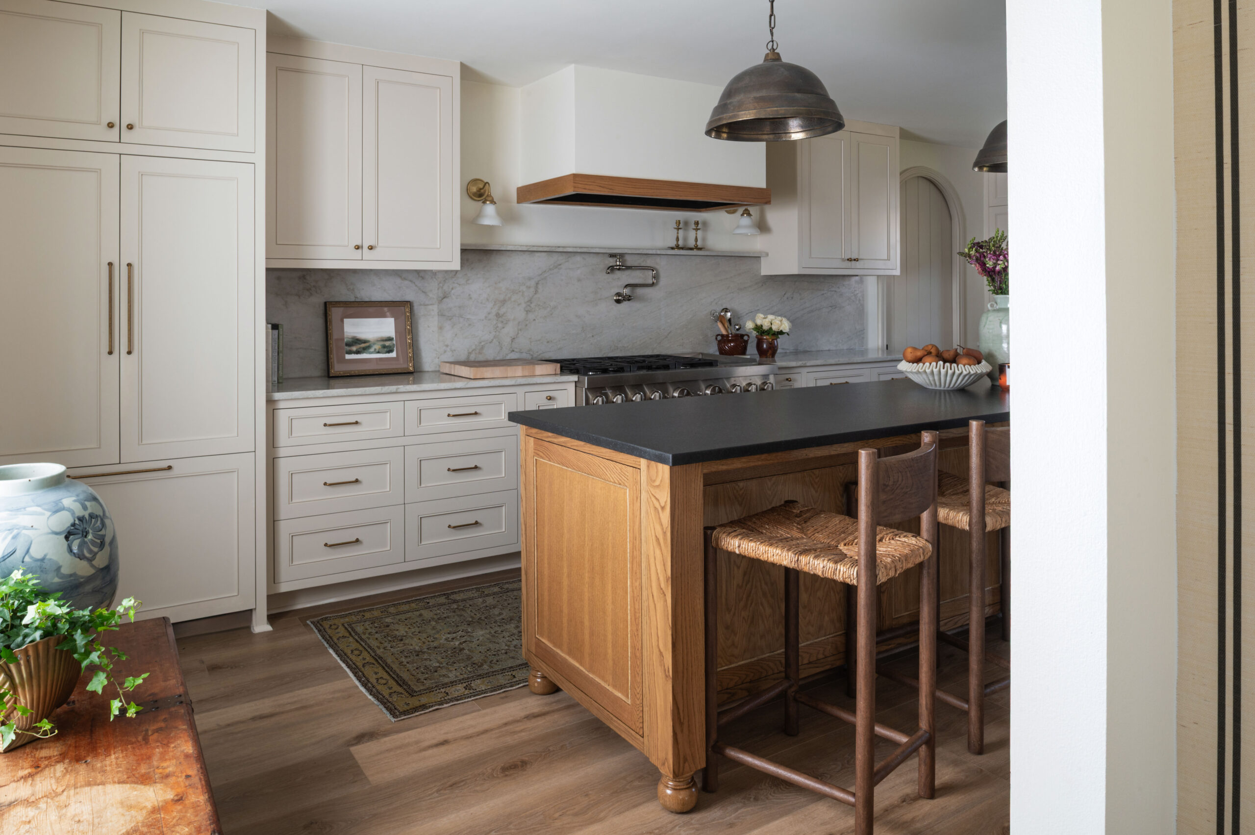 A cozy and timeless kitchen design featuring a black countertop island with oak paneling, woven bar stools, warm brass lighting, and creamy cabinetry—perfect for studying how to photograph interior spaces that emphasize texture and warmth.