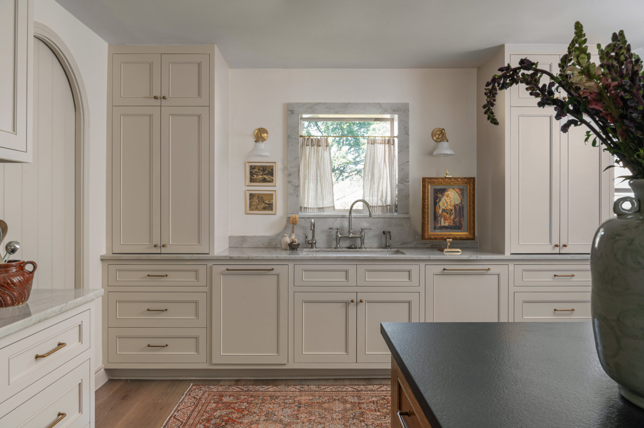 Elegant kitchen sink area with marble countertops, creamy shaker cabinets, antique brass hardware, and a framed art piece near a small window—an inspiring example for how to photograph interior spaces with symmetry and soft natural light.


