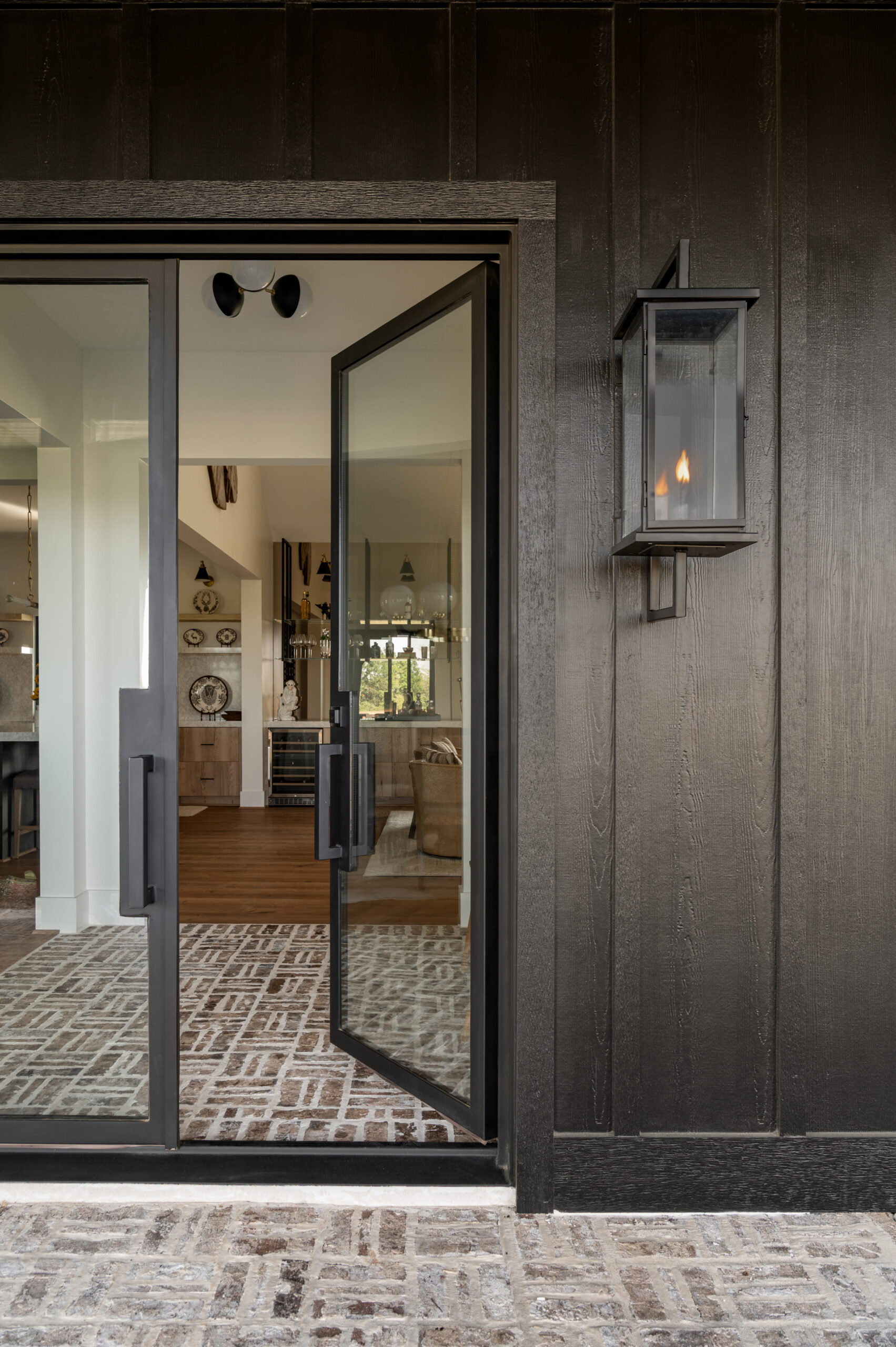 A moody exterior entry featuring black siding, glass double doors, and a lantern sconce, offering a peek into a layered interior full of inviting textures and tones.