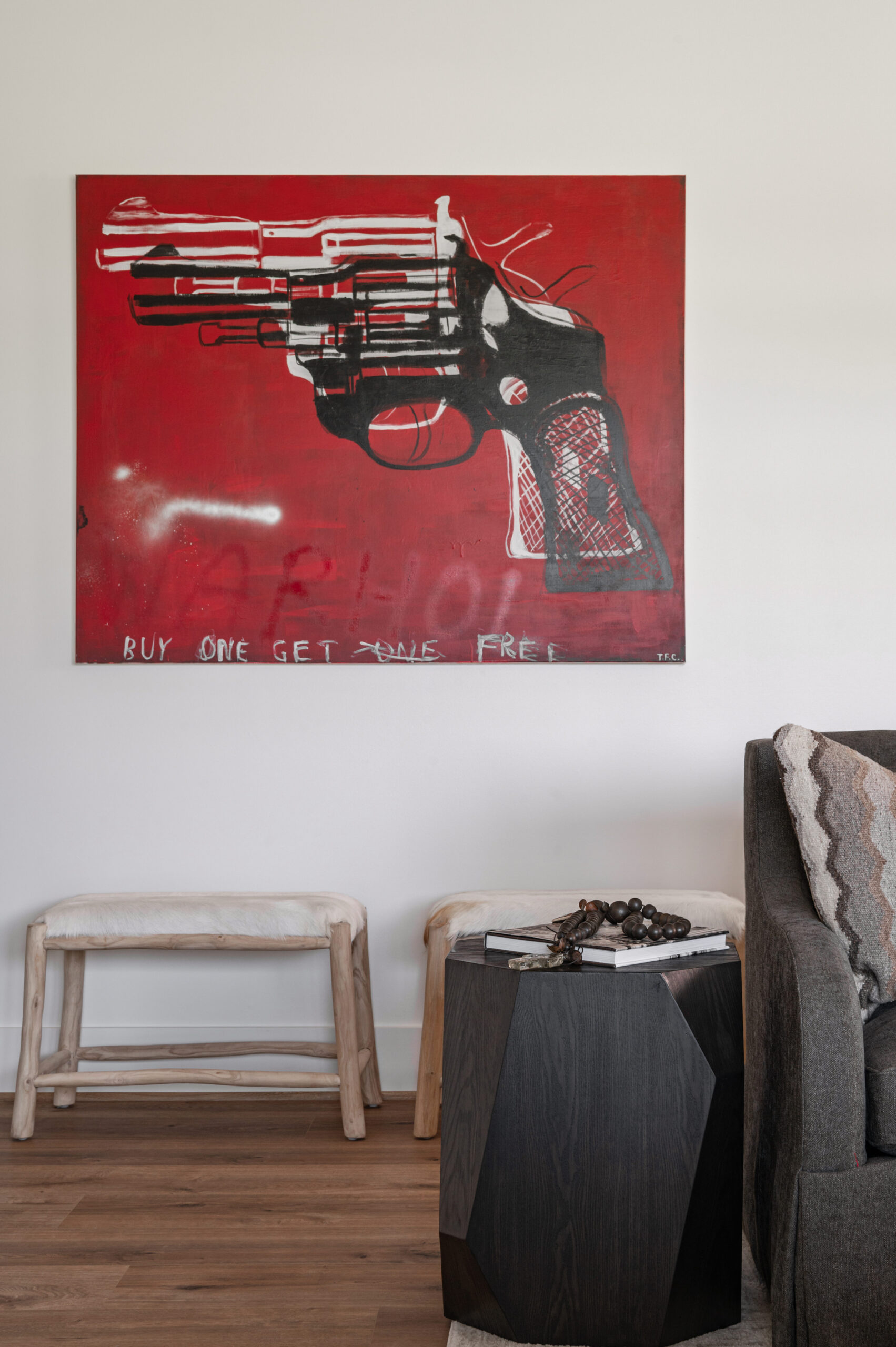  An edgy art piece of a revolver with bold red background and text, displayed above rustic benches and a geometric wood table with books and beads, creating a bold statement corner.