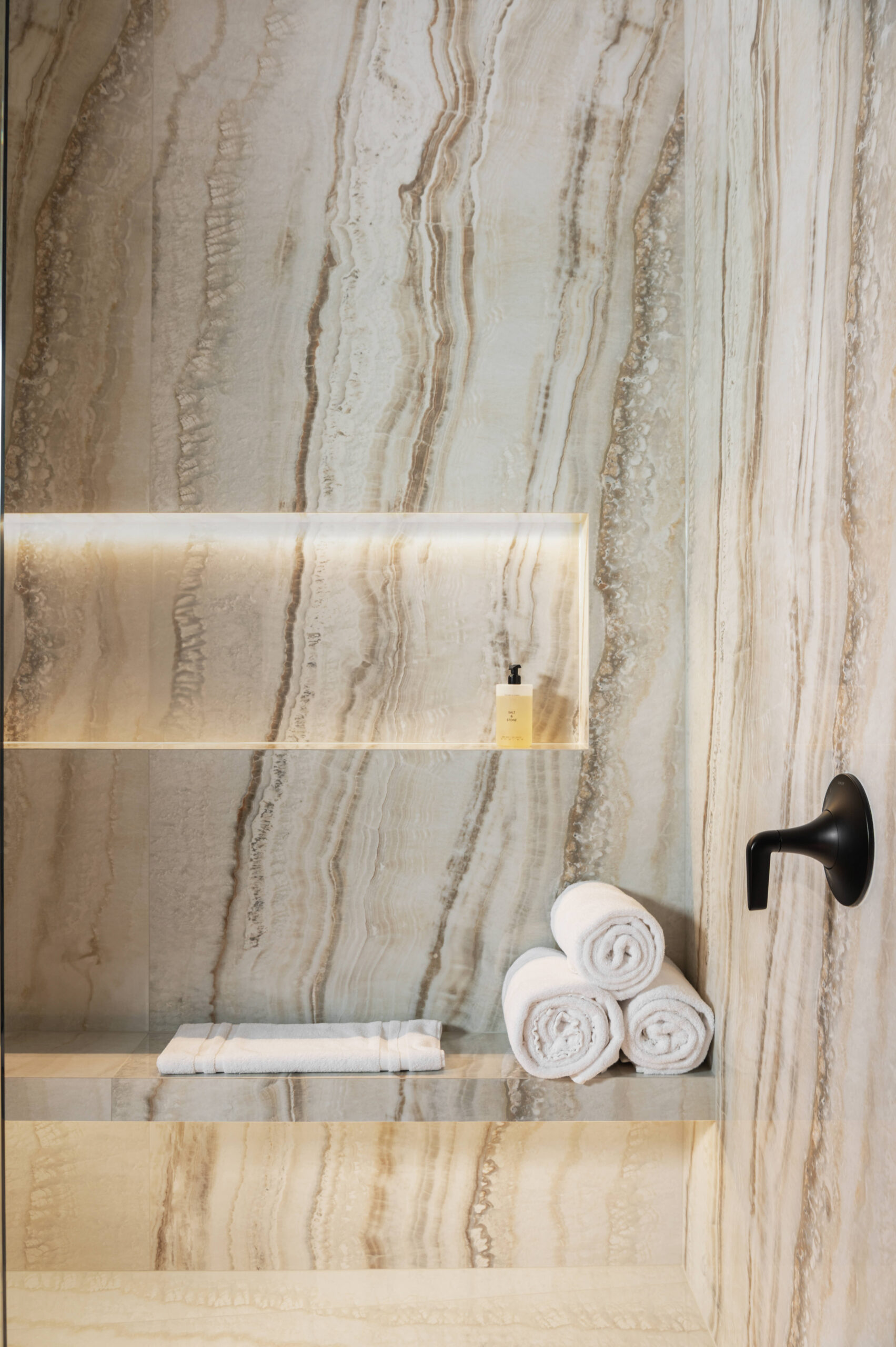 A luxurious walk-in shower with natural stone veining, built-in niches, rolled towels, and minimal matte black fixtures for a clean, spa-like ambiance