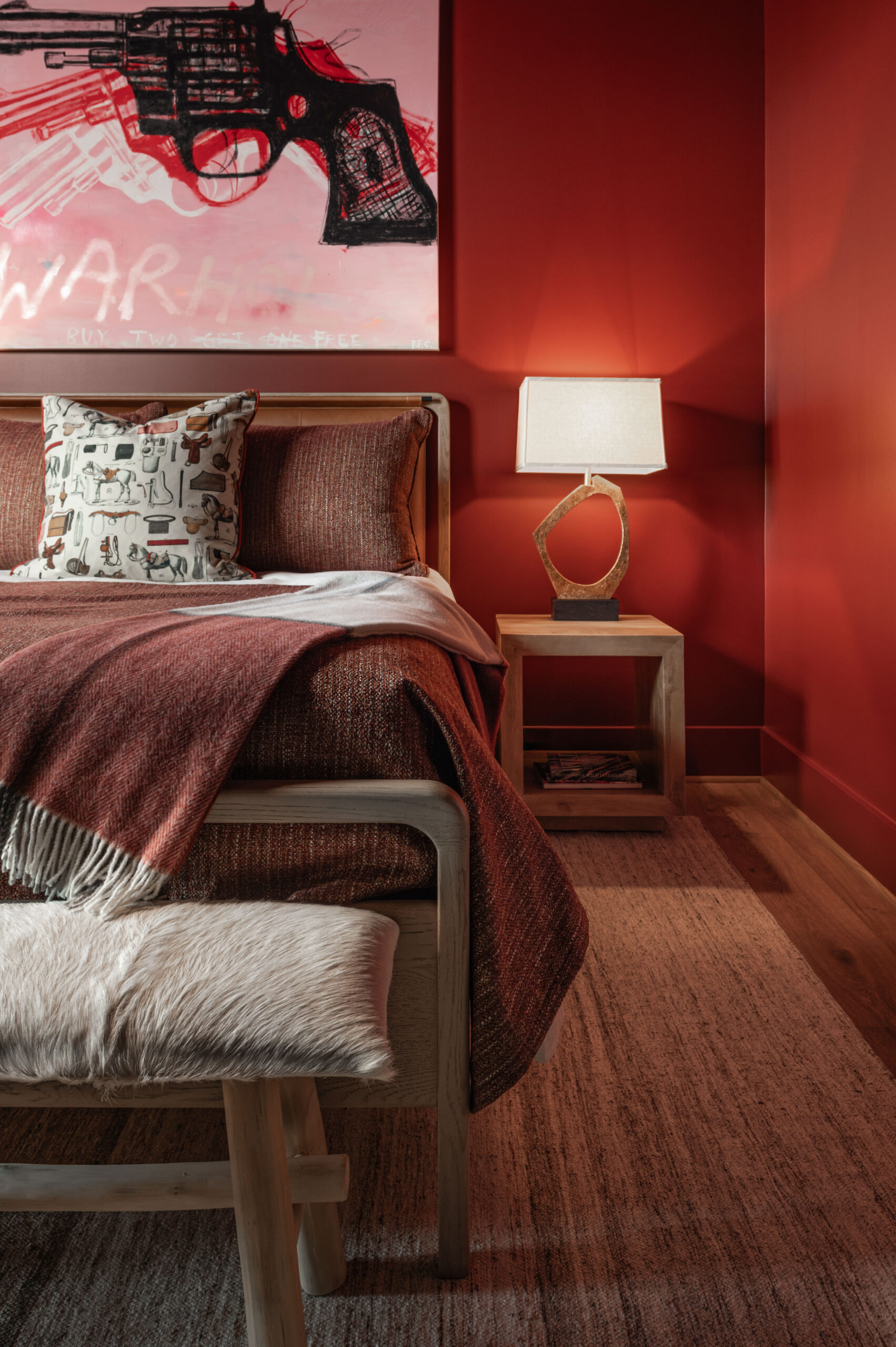 Visual storytelling photography of A neatly made bed with brown bedding, a nightstand with a lamp, a bench with a fur cushion, and a painting of a red gun on the wall in a red-toned bedroom