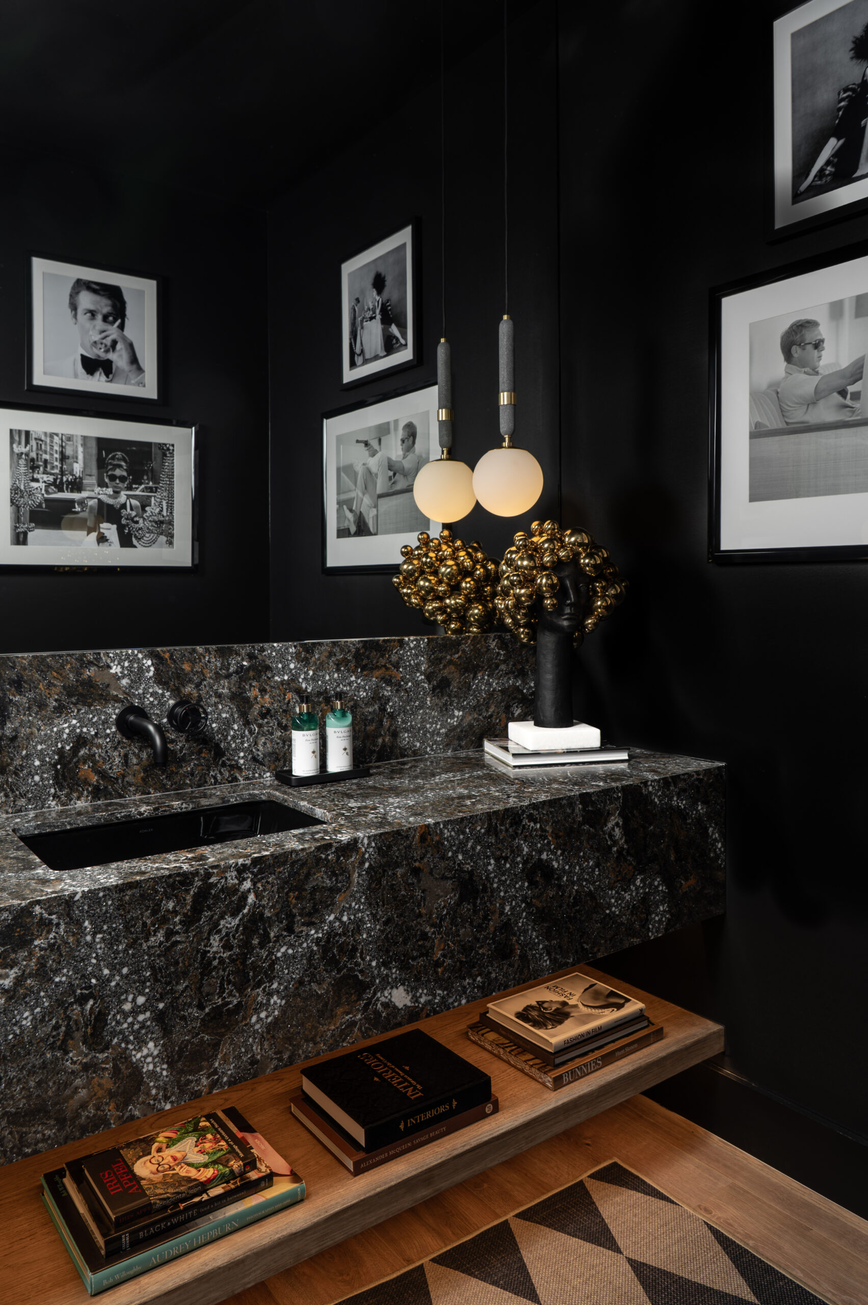 Visual storytelling photography of Modern bathroom with black walls, marble countertop, two globe pendant lights, framed black-and-white photos, and books on a wooden shelf beneath the sink