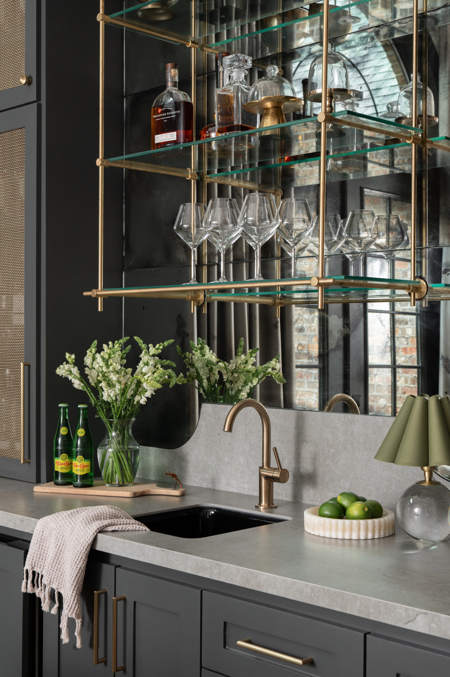 Close-up of a bar area showcasing a brass faucet, under-mount sink, fresh flowers, and drinks beneath glass shelving styled with barware and a mirror backsplash—perfect for a modern interior design portfolio.