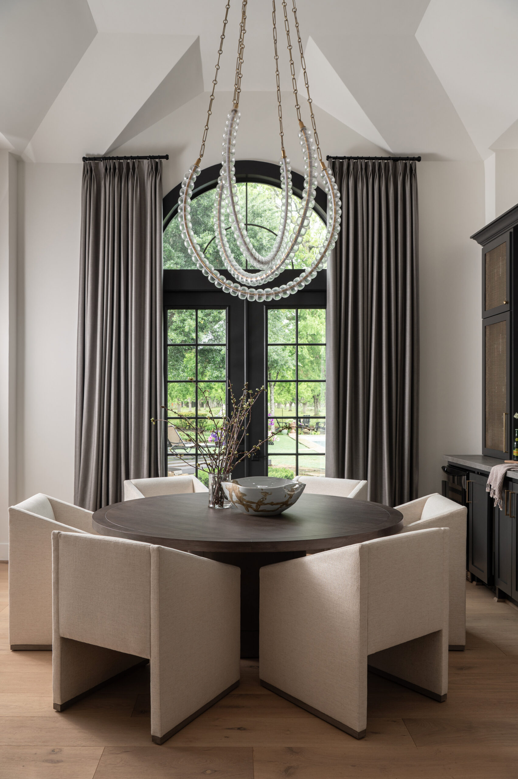 A softly lit dining room featuring a round dark wood table, sculptural cream chairs, and a modern beaded chandelier framed by tall gray curtains and black-trimmed windows.