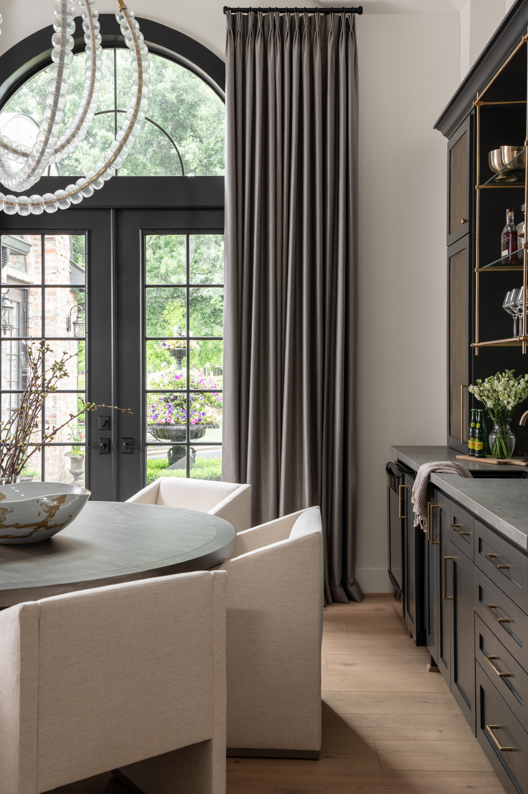 A softly lit dining room featuring a round dark wood table, sculptural cream chairs, and a modern beaded chandelier framed by tall gray curtains and black-trimmed windows.