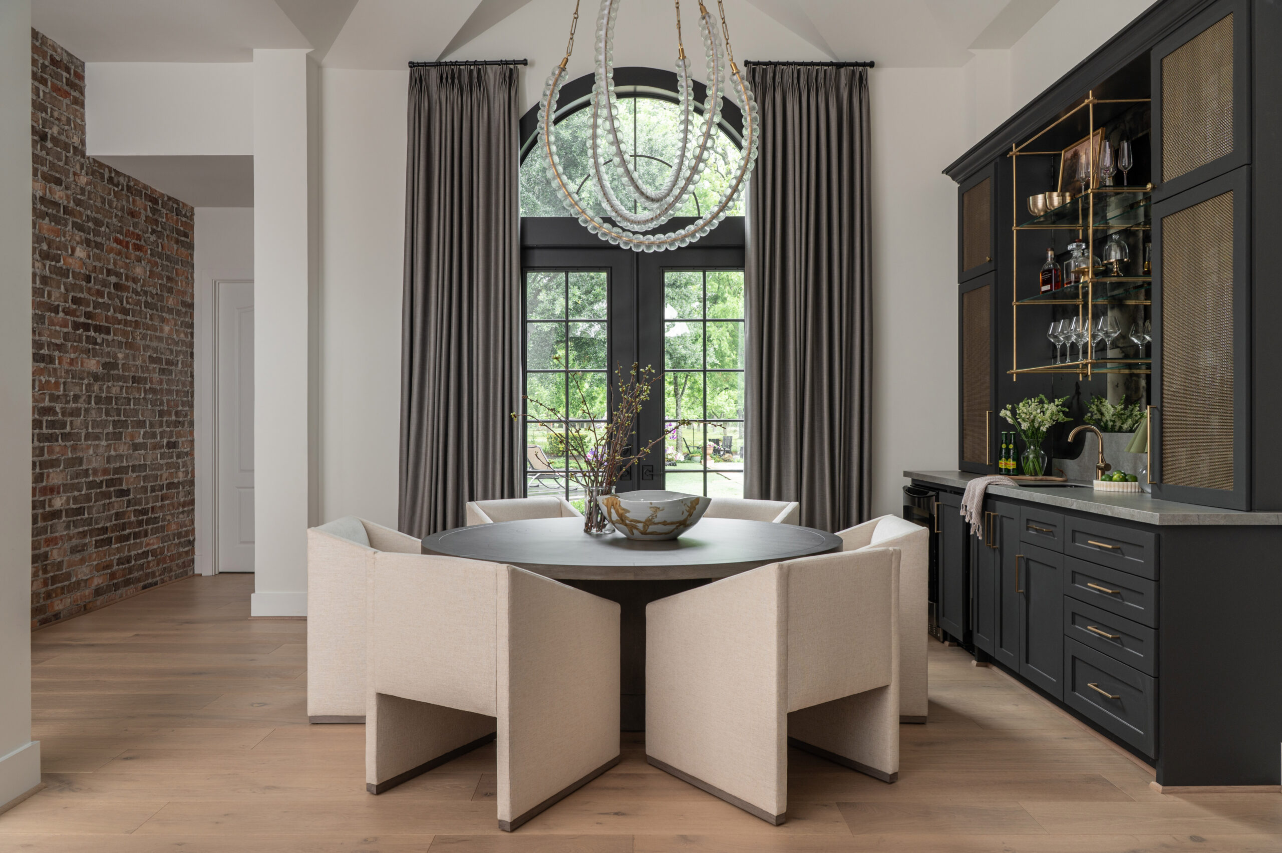 A softly lit dining room featuring a round dark wood table, sculptural cream chairs, and a modern beaded chandelier framed by tall gray curtains and black-trimmed windows.