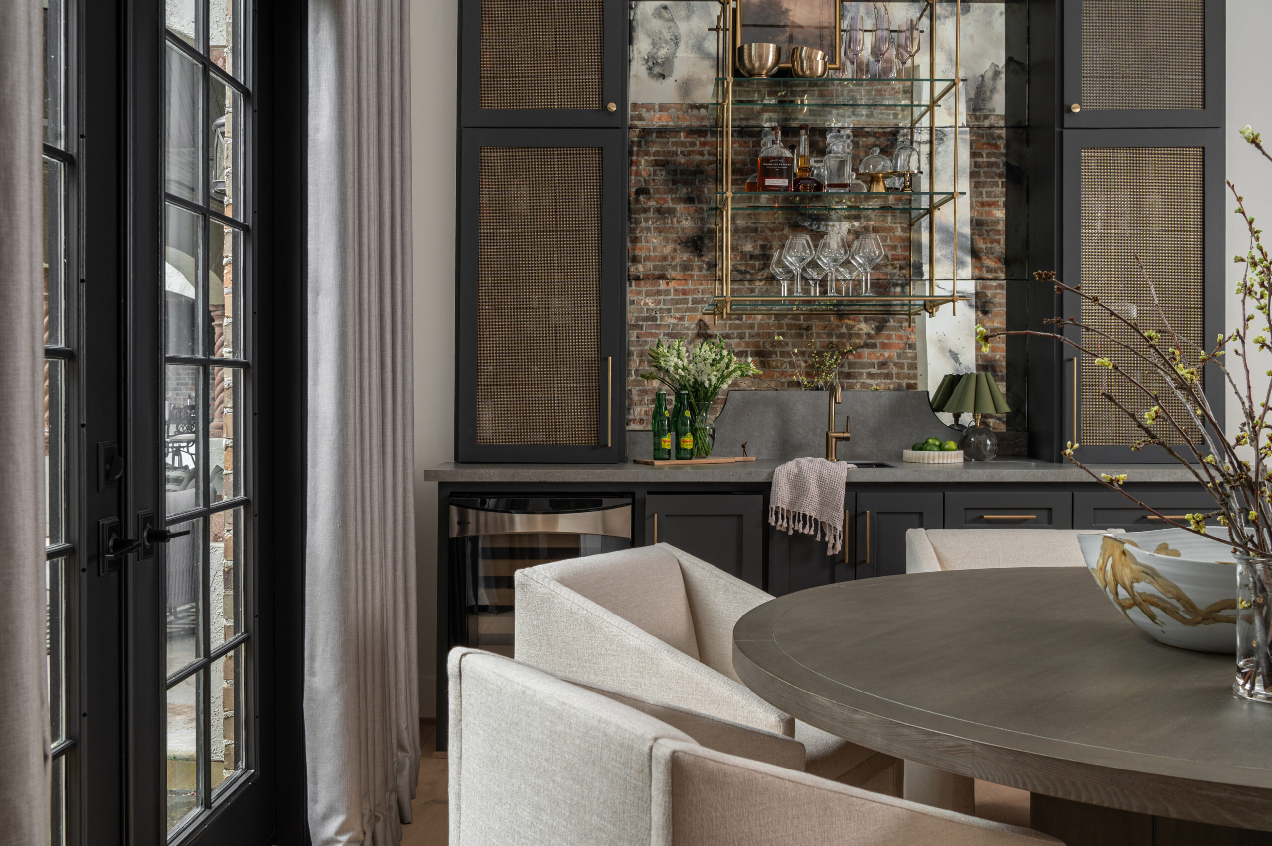 A softly lit dining room featuring a round dark wood table, sculptural cream chairs, and a modern beaded chandelier framed by tall gray curtains and black-trimmed windows.