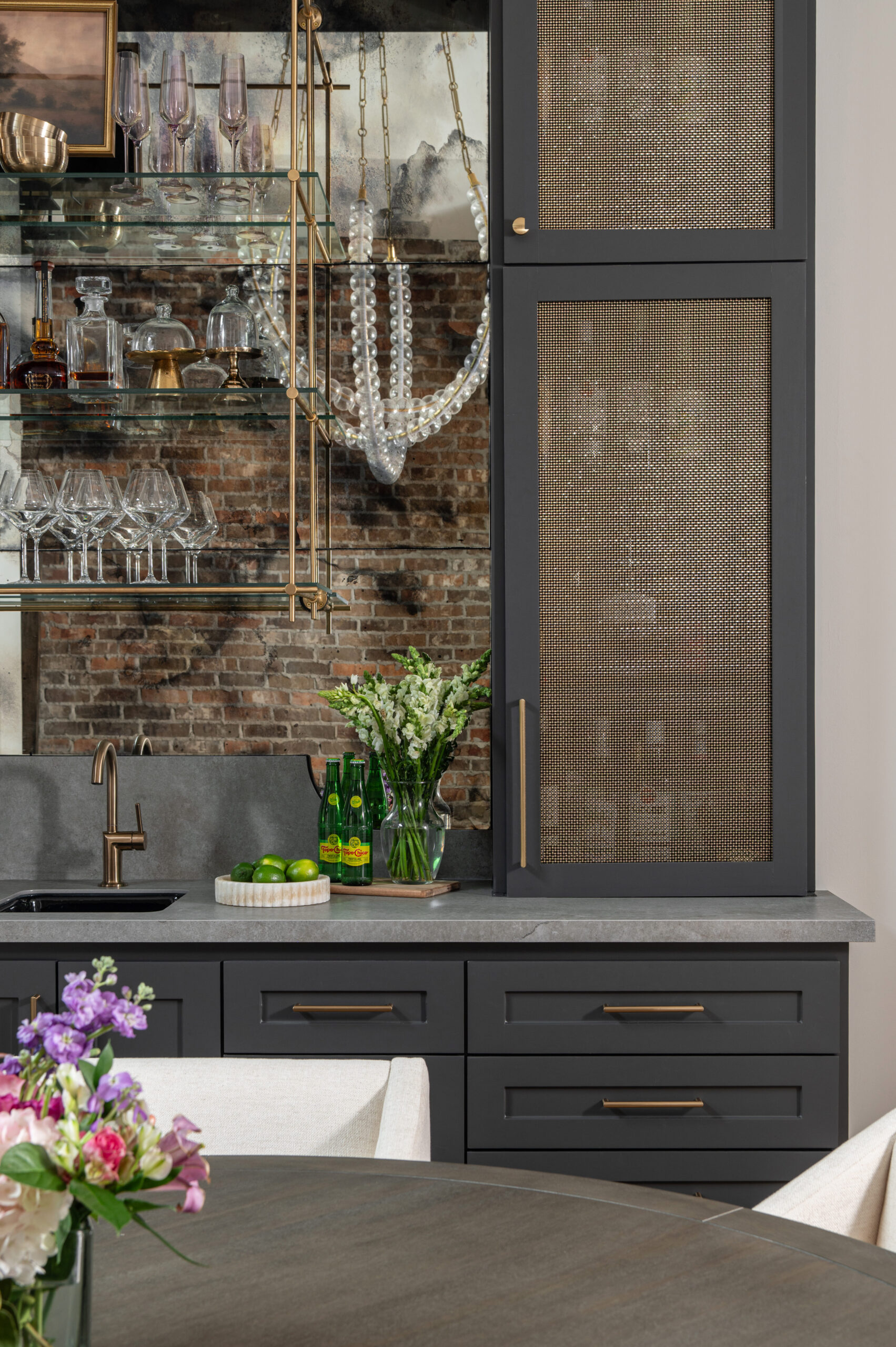 A stylish home bar featuring dark cabinetry, a matte gray countertop, and open brass-framed glass shelving against a distressed mirrored backsplash, styled with glassware, florals, and limes for a lived-in touch