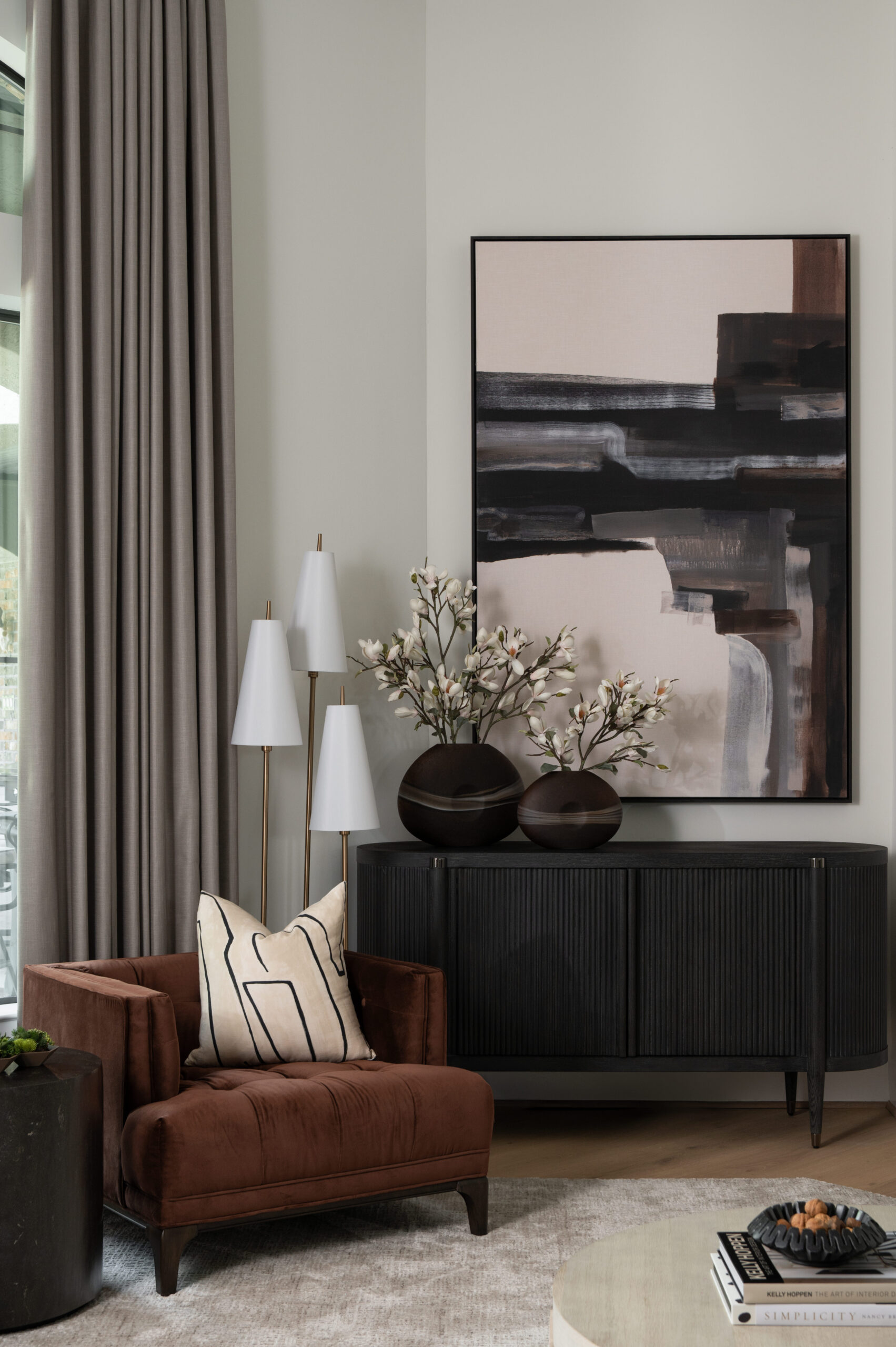 A cozy reading nook in a neutral-toned living room, styled with a rust velvet chair, layered pillows, a white floor lamp trio, and leafy greenery for a warm, inviting space.