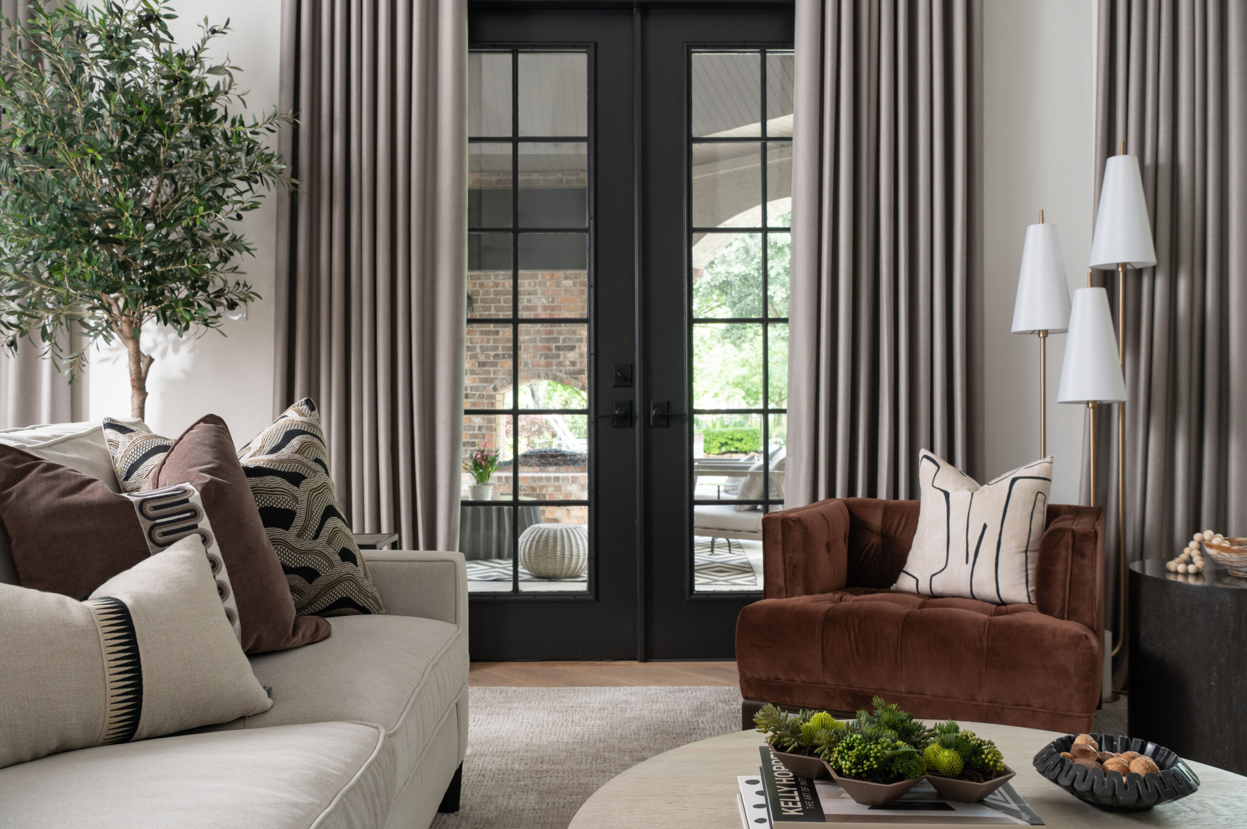 A cozy reading nook in a neutral-toned living room, styled with a rust velvet chair, layered pillows, a white floor lamp trio, and leafy greenery for a warm, inviting space.