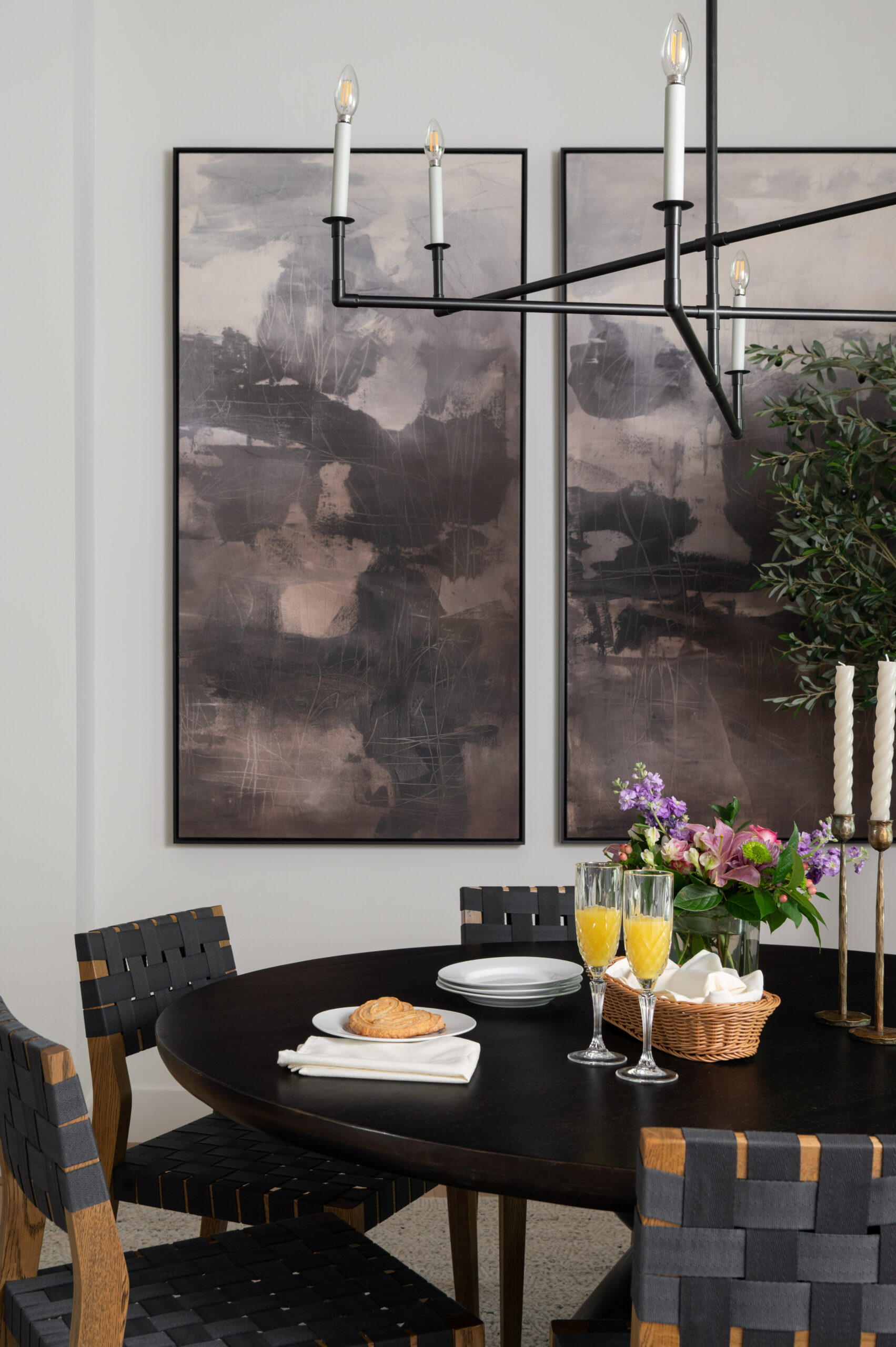 Contemporary dining room with a round black table, six woven black and wood chairs, and an oversized olive tree under a minimalist black chandelier, styled for a portfolio