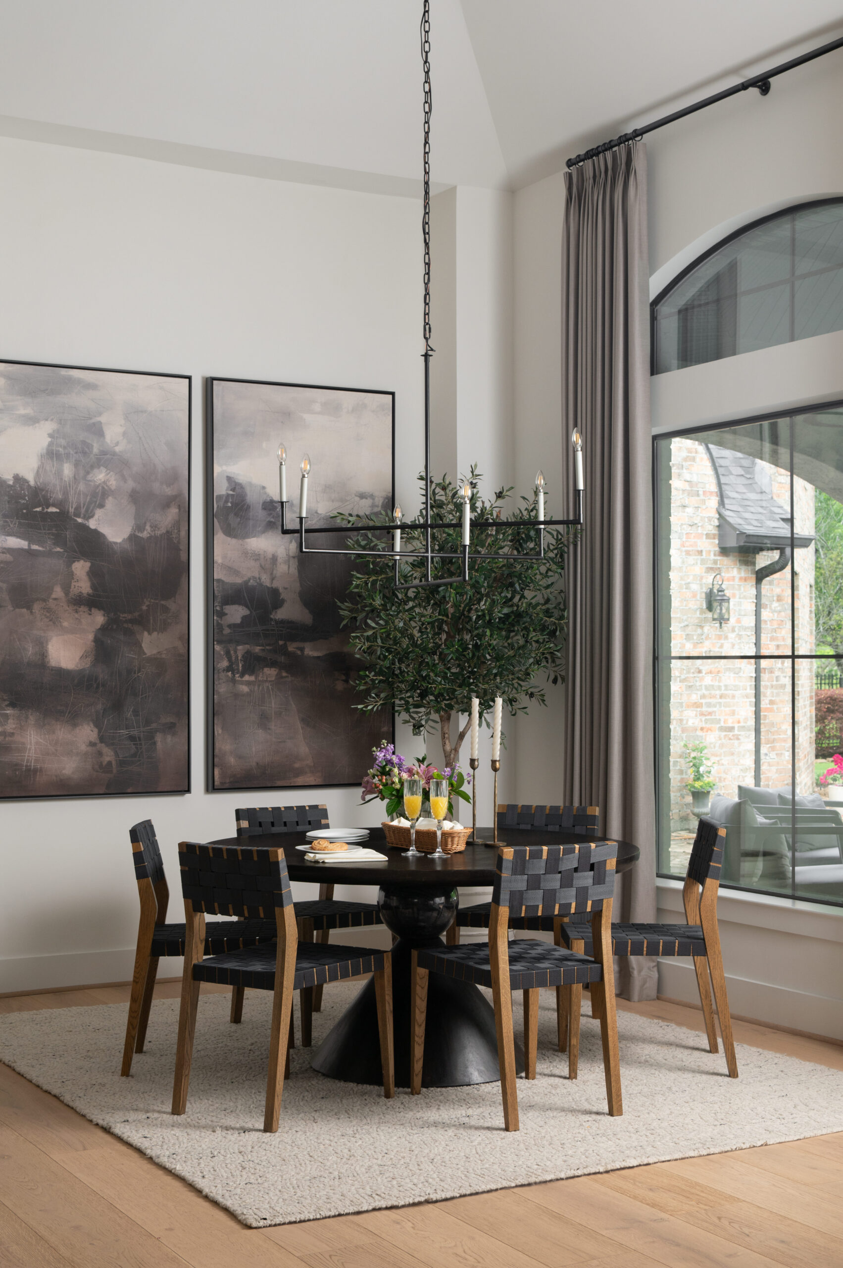 Contemporary dining room with a round black table, six woven black and wood chairs, and an oversized olive tree under a minimalist black chandelier, styled for a portfolio