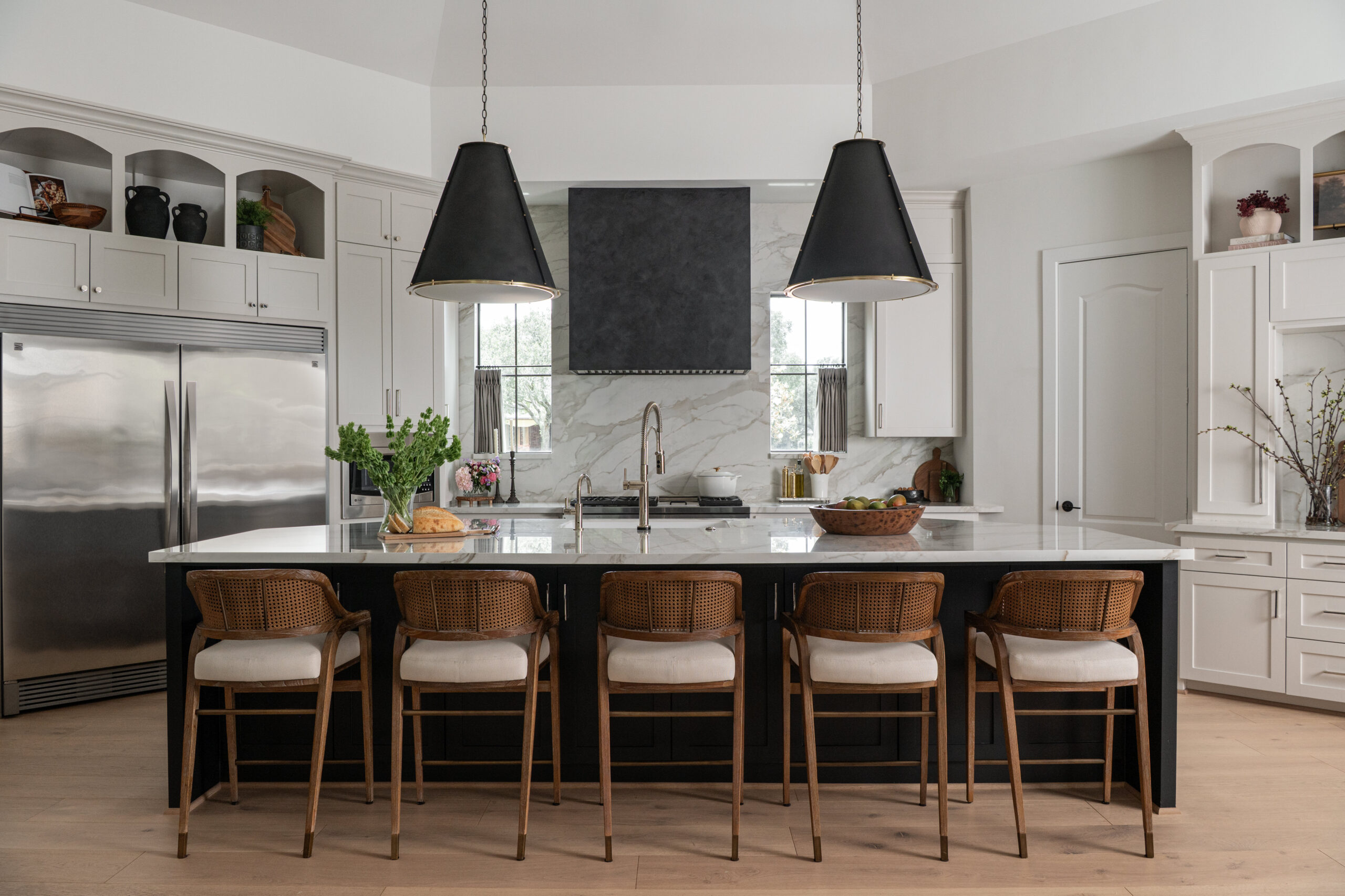 A spacious transitional kitchen featuring a dark central island with wooden bar stools, dramatic black pendant lighting, and white cabinetry accented by veined stone walls—ideal for a portfolio for interior design.