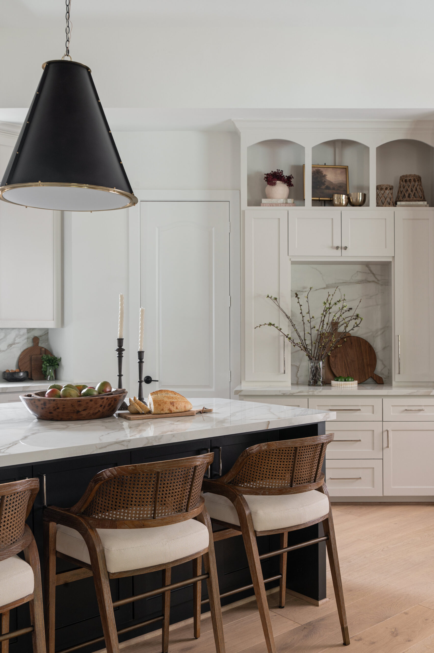 A spacious transitional kitchen featuring a dark central island with wooden bar stools, dramatic black pendant lighting, and white cabinetry accented by veined stone walls—ideal for a portfolio for interior design.