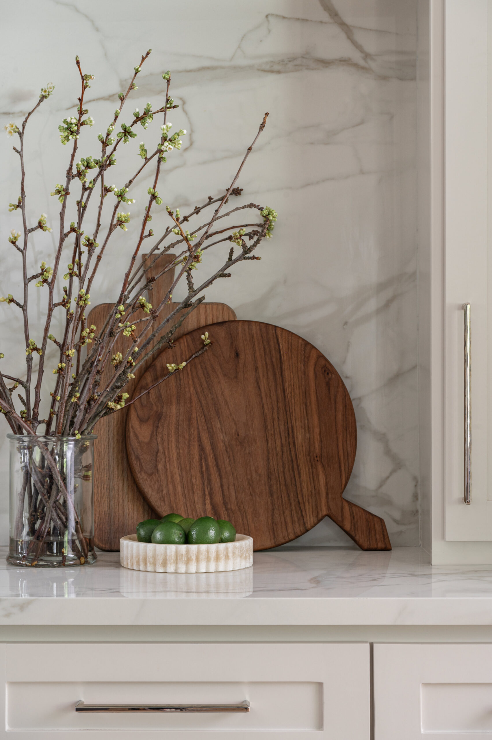 A close-up of a marble countertop vignette, featuring layered wooden cutting boards, a bowl of limes, and budding branches in a glass vase for a natural, textural styling moment.