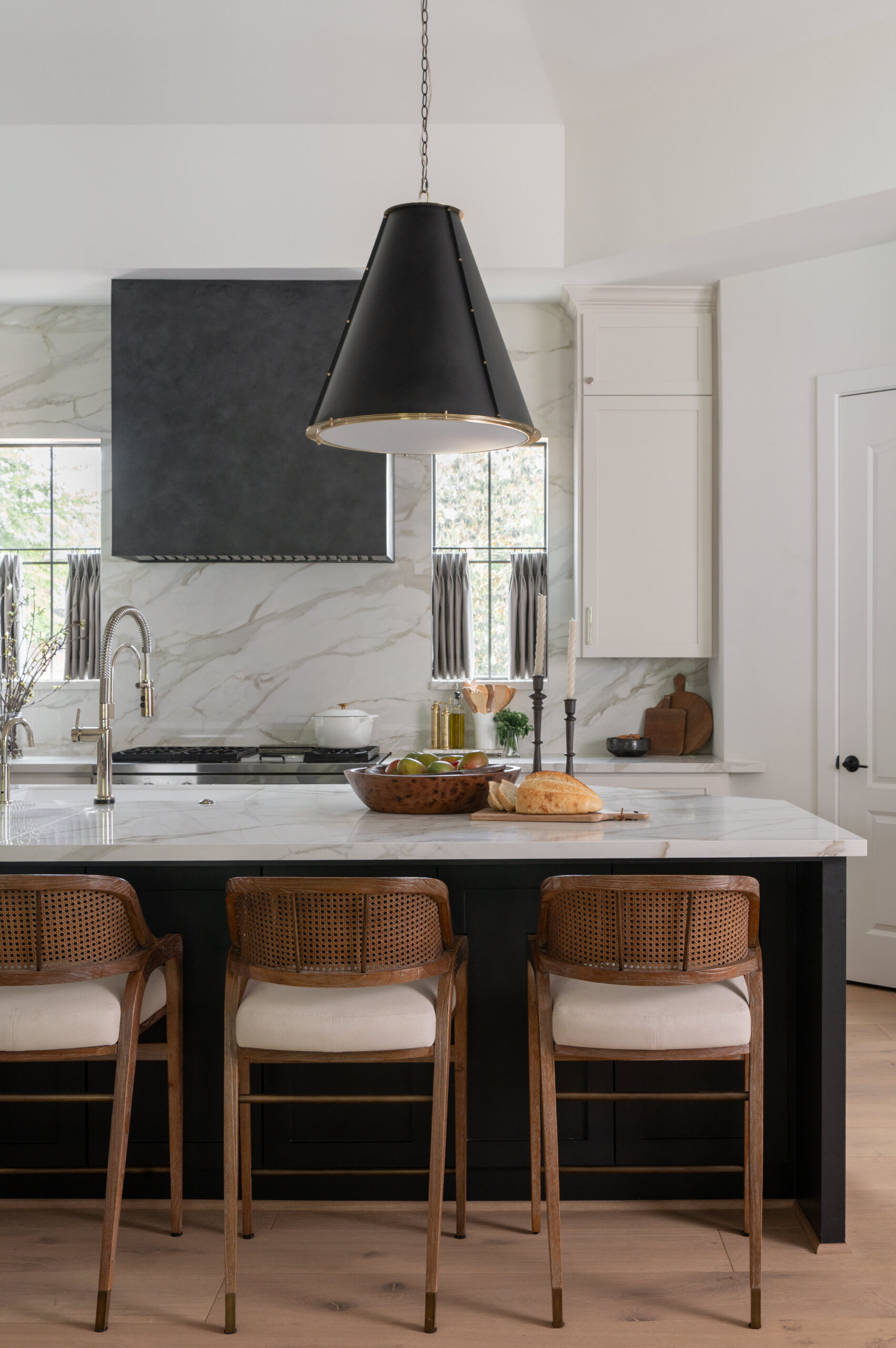A marble kitchen island surrounded by rattan and cushion bar stools, with a sleek black range hood and natural light pouring through window panels behind it.