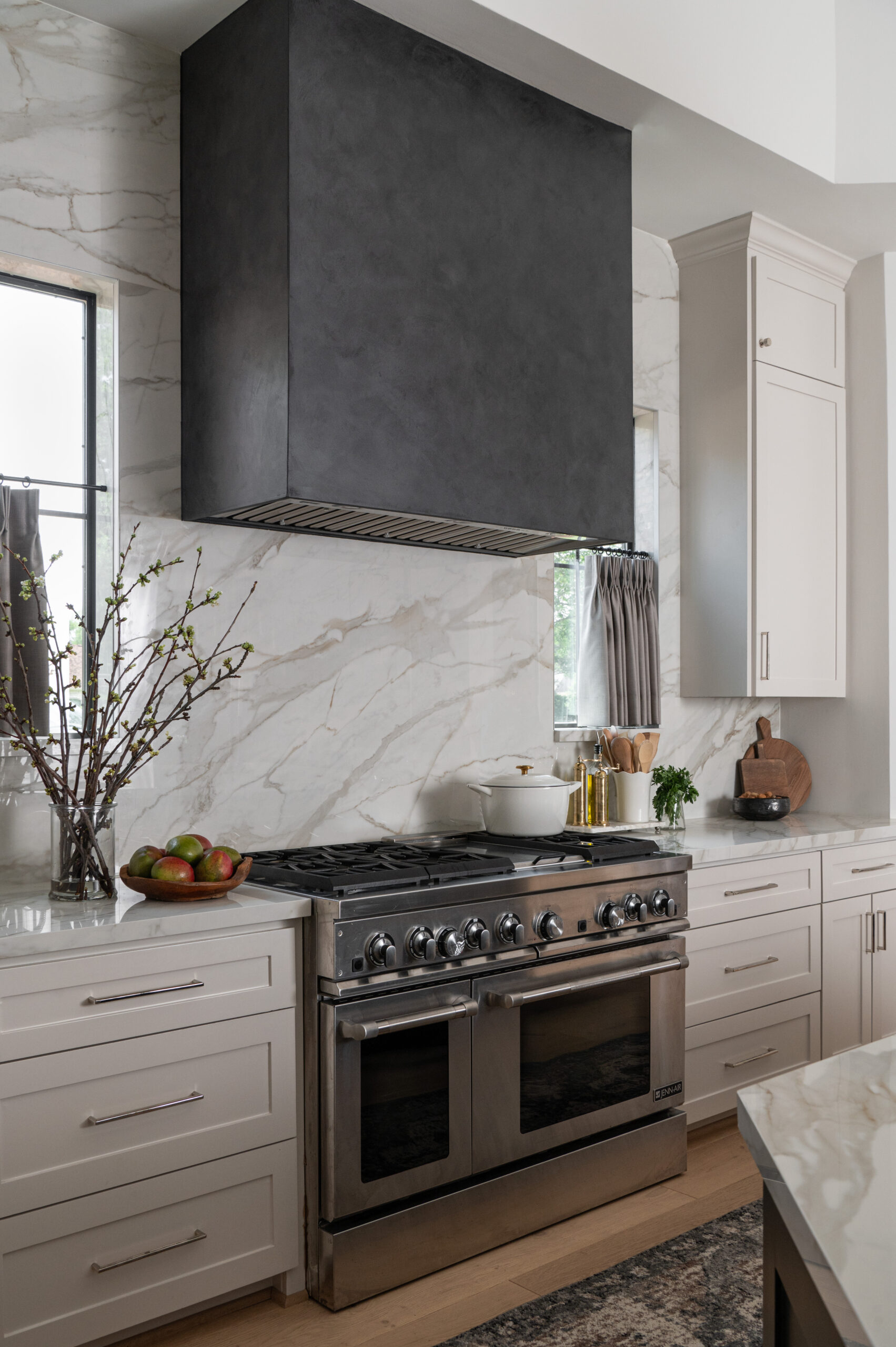 A professional-grade stainless steel range under a dark plaster vent hood, with a marble backsplash and bright cabinetry—highlighting the heart of the home for a portfolio for interior design.