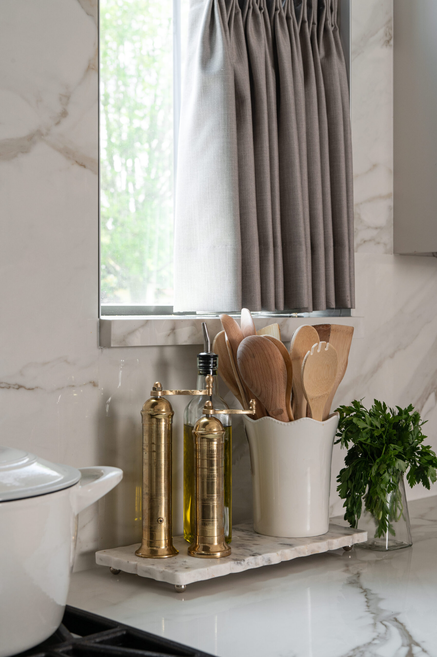 A detail shot of a kitchen counter styled with gold oil bottles, wooden utensils in a ceramic crock, and fresh herbs beside a window with pleated gray curtains.