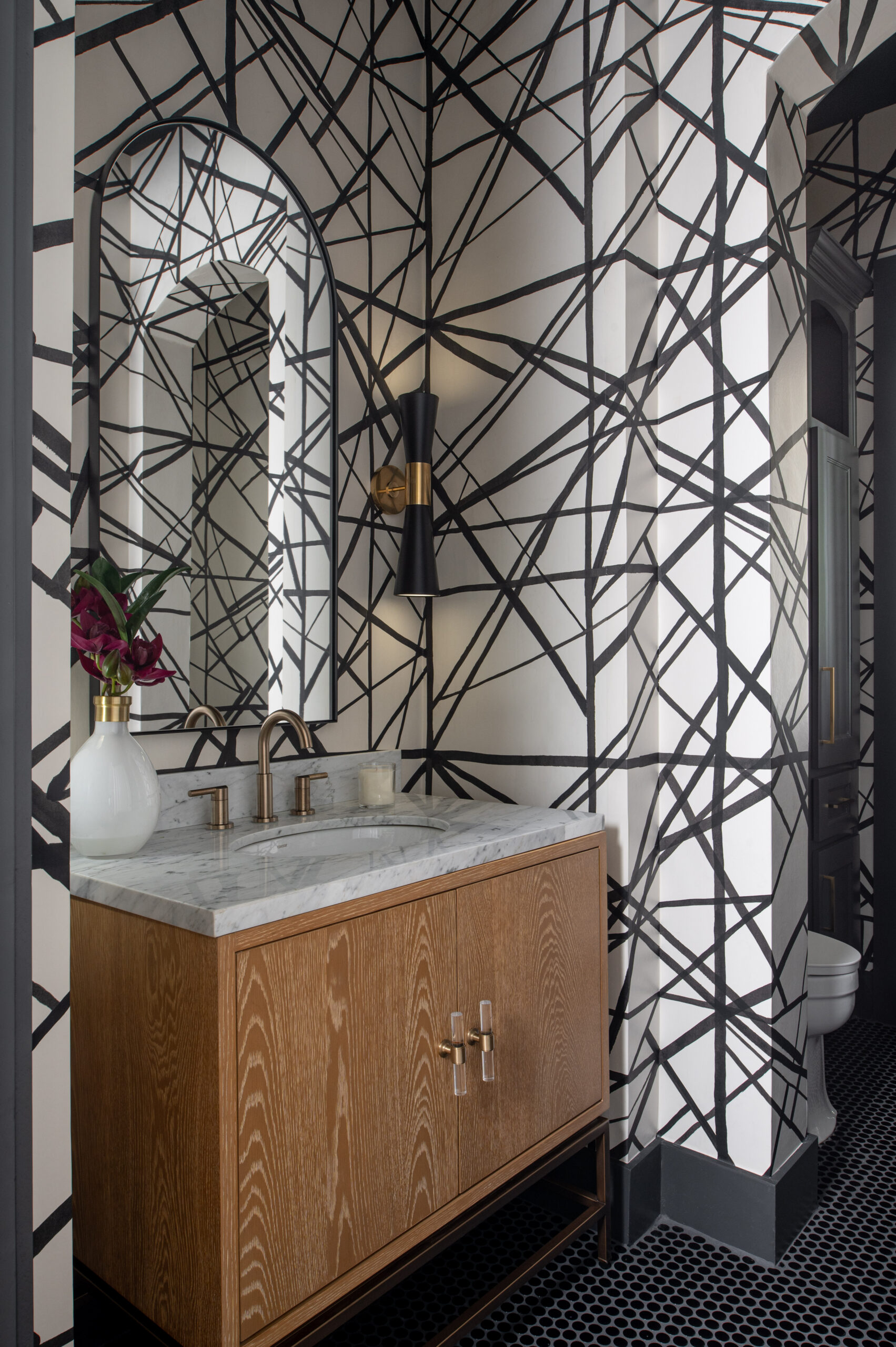 A bold powder bathroom featuring graphic black-and-white wallpaper, a wood vanity with marble countertop, arched mirror, and sculptural sconces—a striking example of contrast and texture ideal for a portfolio