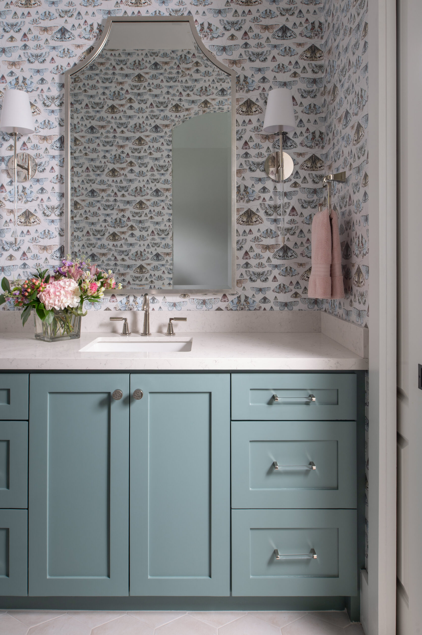 Powder room with soft blue cabinetry, a crisp white quartz countertop, scalloped double mirrors, and whimsical mountain wallpaper, styled with fresh florals.