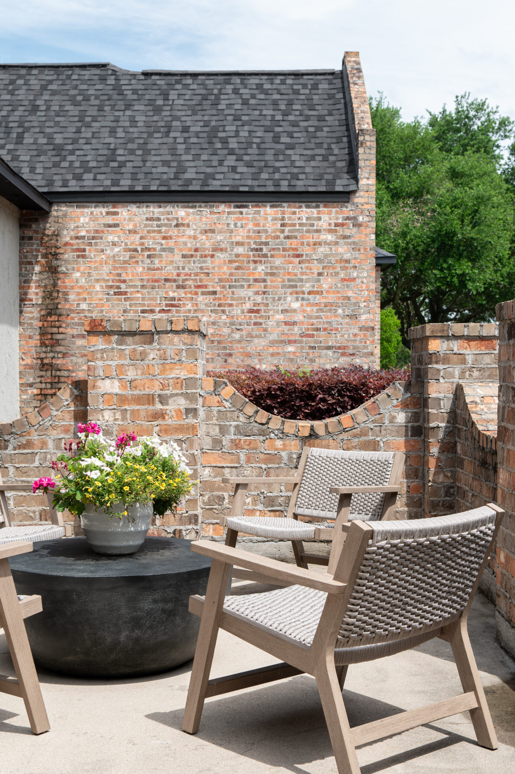 Cozy outdoor patio space featuring a round black coffee table, four woven wood chairs, and a stone façade under soft natural light.
