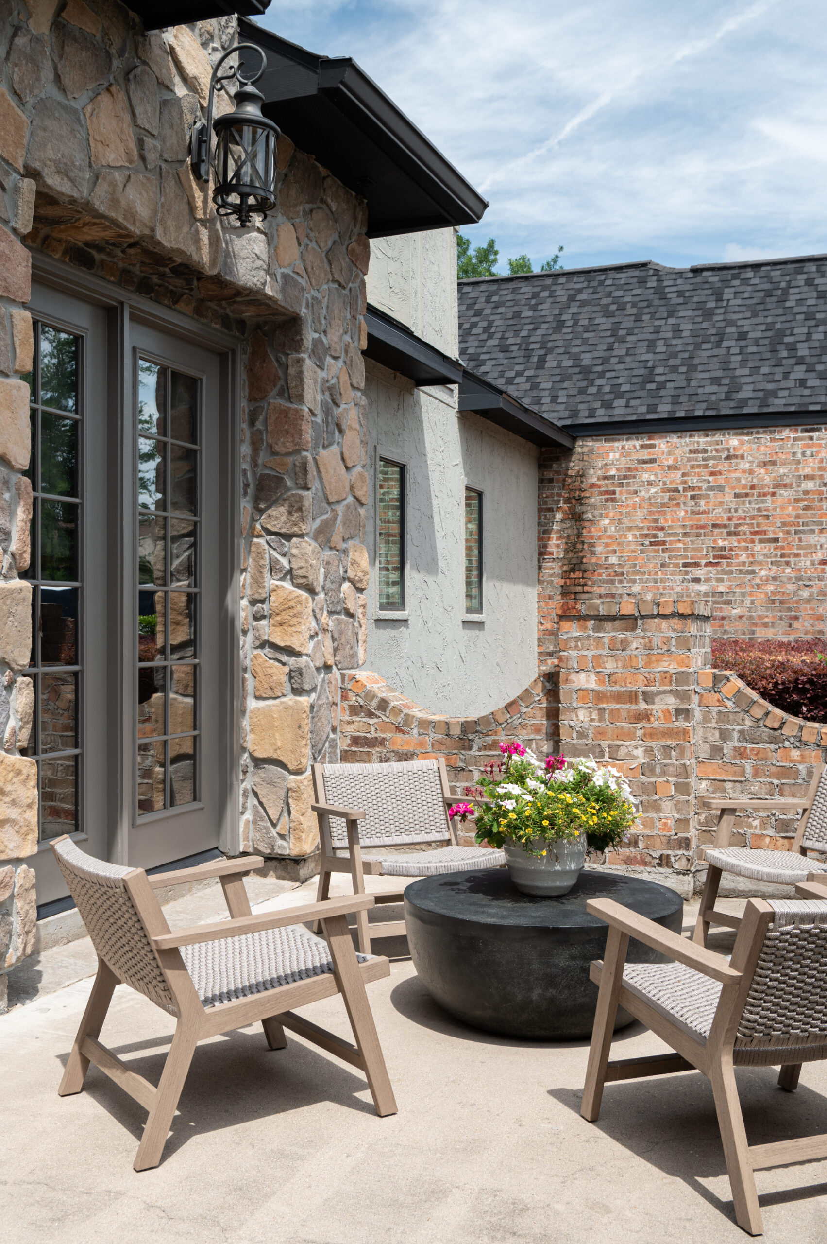 Cozy outdoor patio space featuring a round black coffee table, four woven wood chairs, and a stone façade under soft natural light.