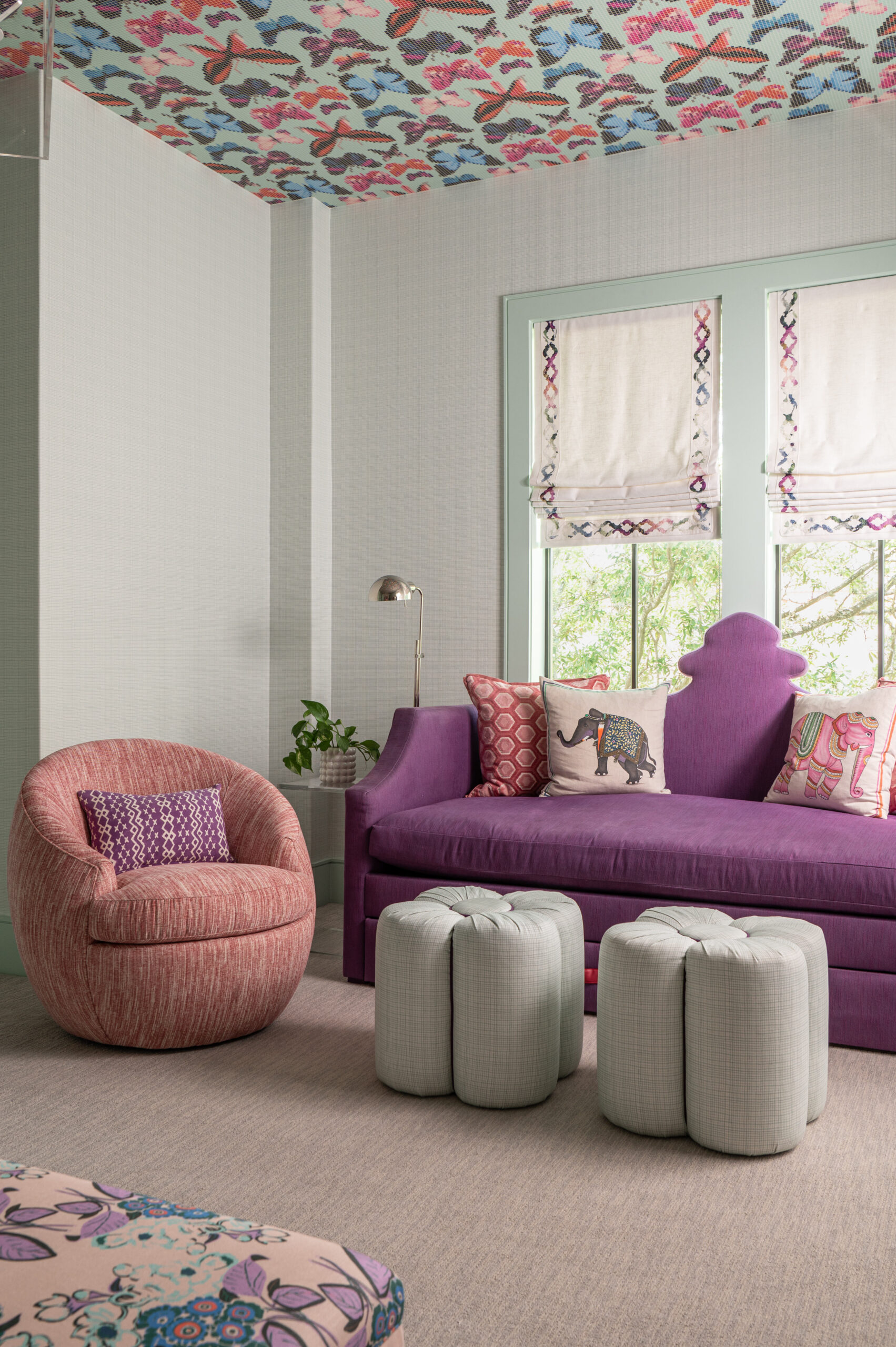 A vibrant tween seating area with a plush lavender daybed, pink chair, and two white ottomans under large windows.