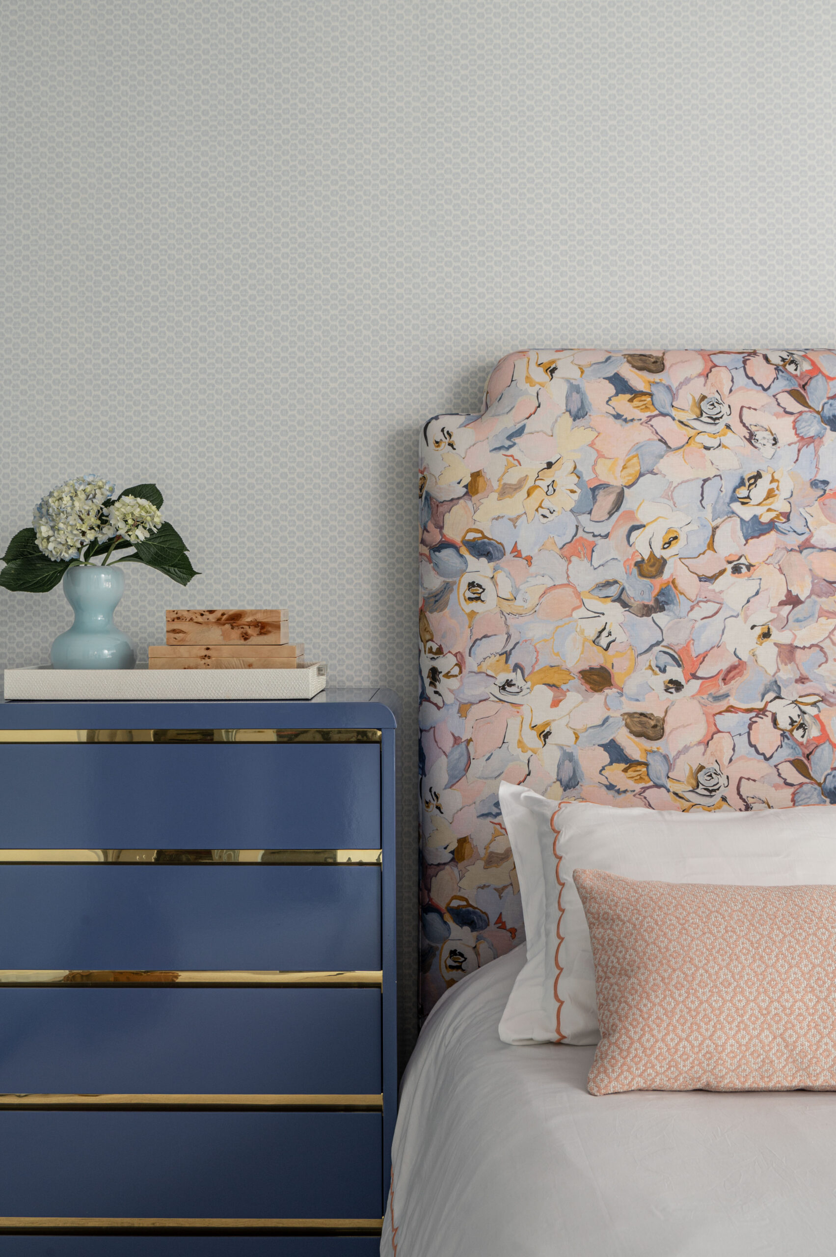 A close-up of a bold floral headboard next to a navy blue nightstand with gold accents and a small flower arrangement.