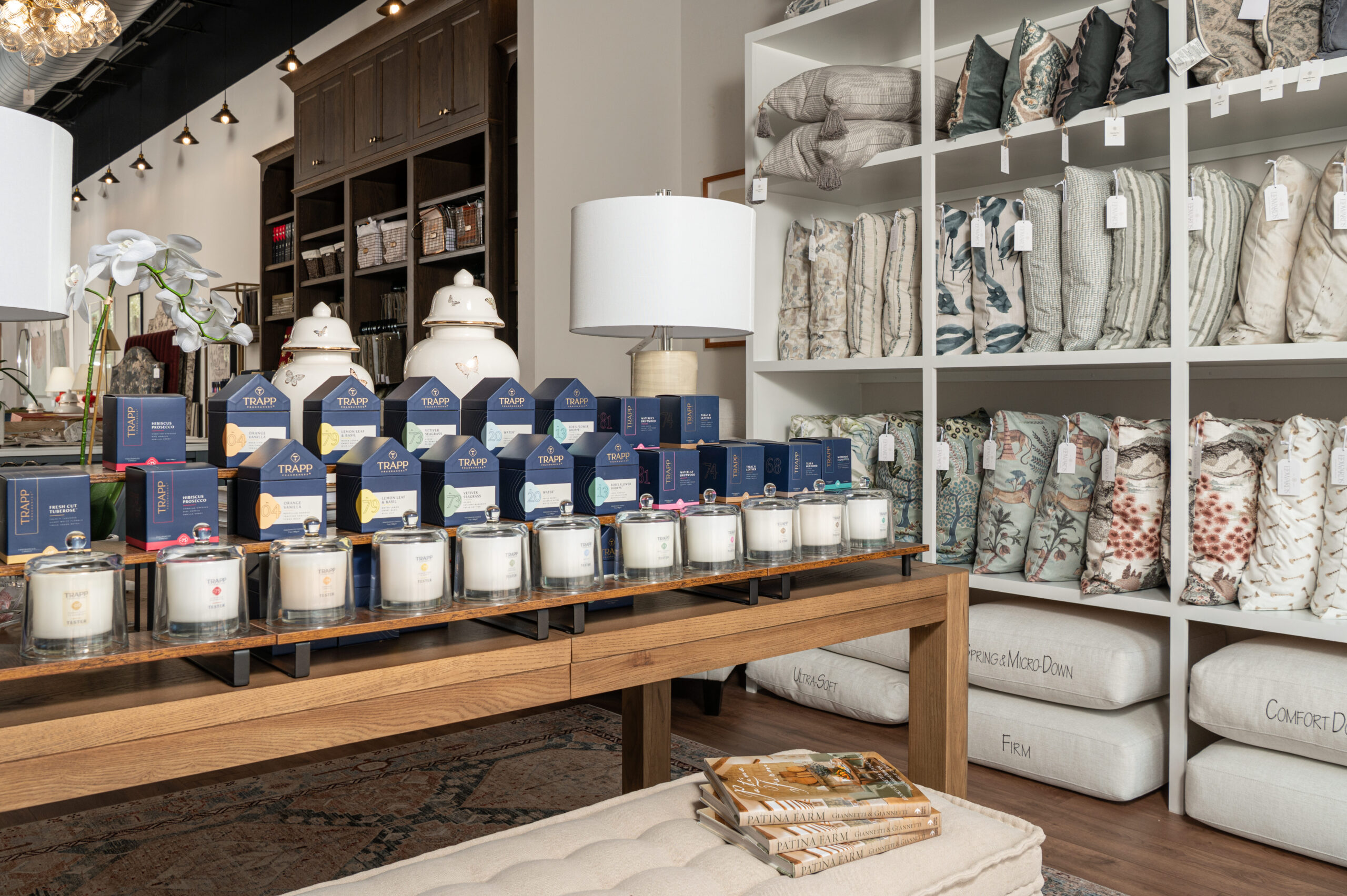 Retail display of Trapp candles, decorative pillows, and luxury bedding in an upscale home goods store, styled with a neutral palette and warm wood tones.