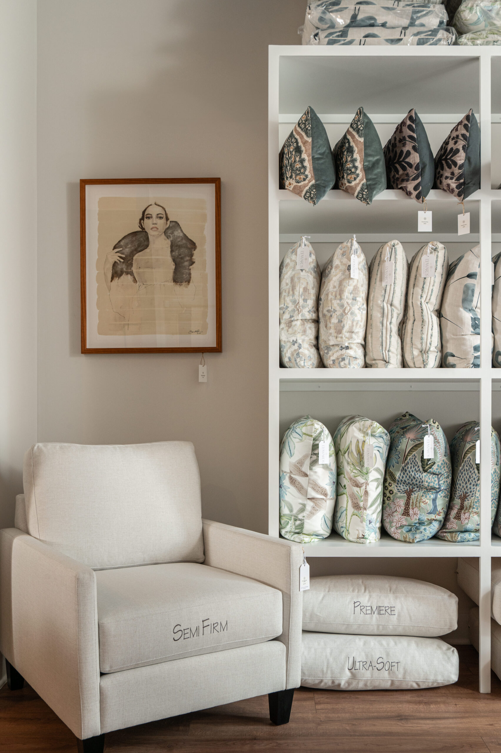 Neutral armchair in a showroom corner next to a built-in shelf filled with decorative pillows and bolsters, showcasing various fabrics, patterns, and fill options.
