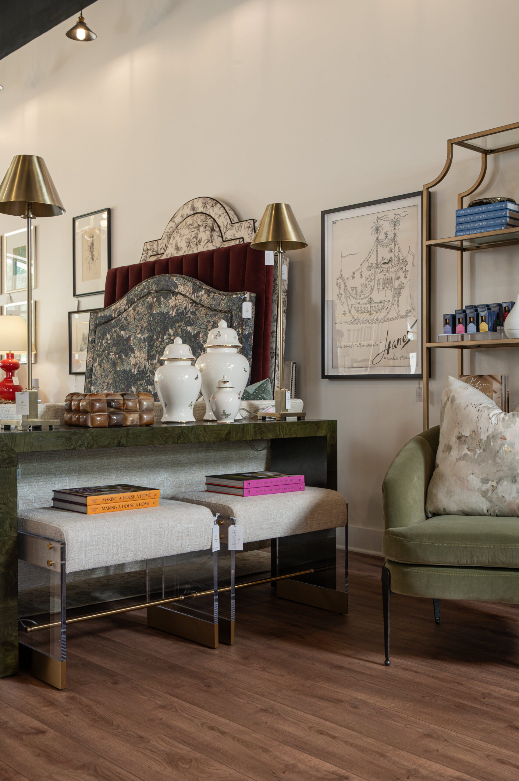 Eclectic showroom vignette featuring a velvet upholstered headboard, chinoiserie art, brass lamps, decorative books, and layered accent benches with acrylic legs.

