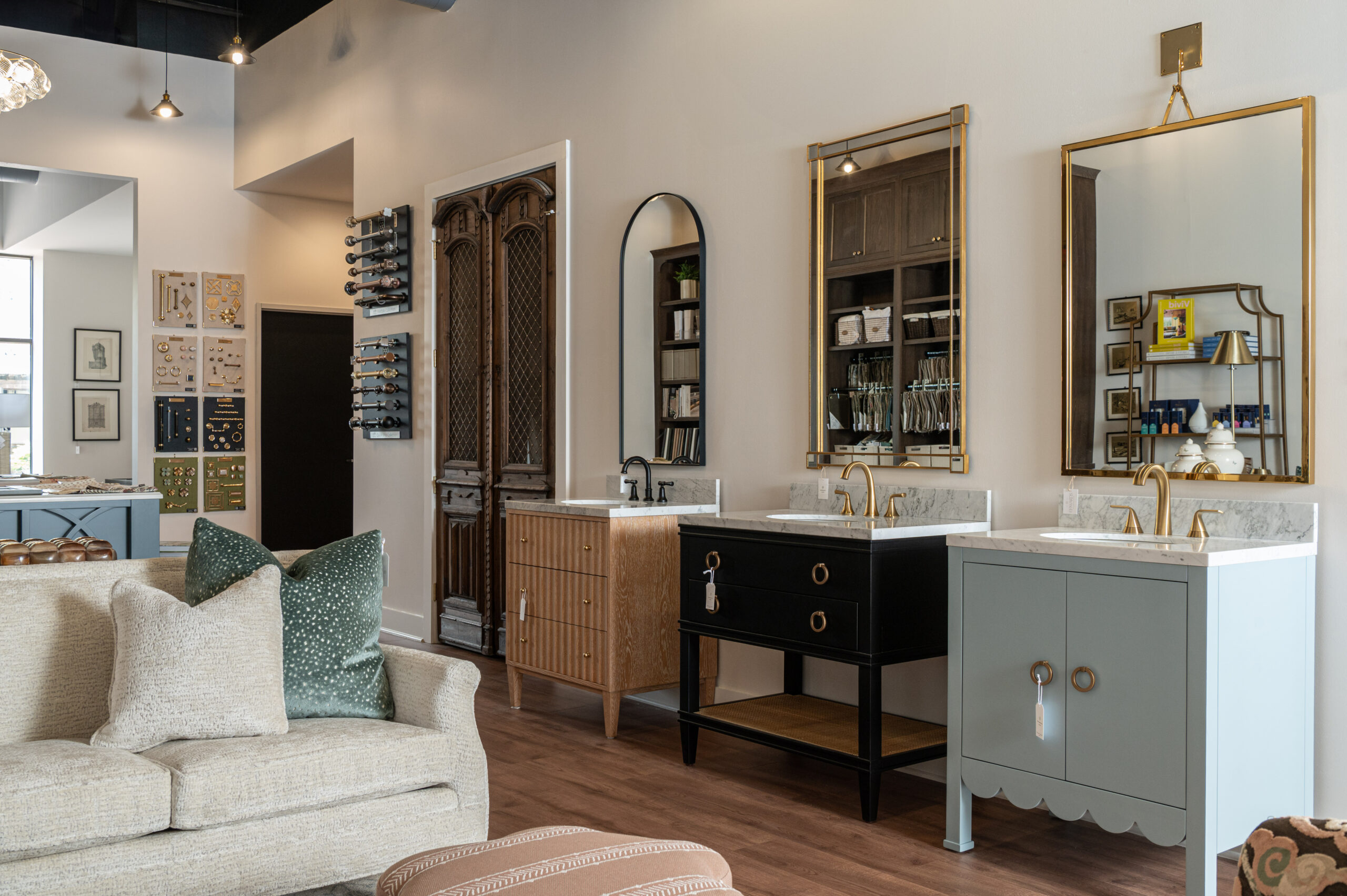 Styled showroom display of bathroom vanities featuring different wood finishes, gold hardware, and marble countertops, surrounded by curated hardware samples and cozy seating.