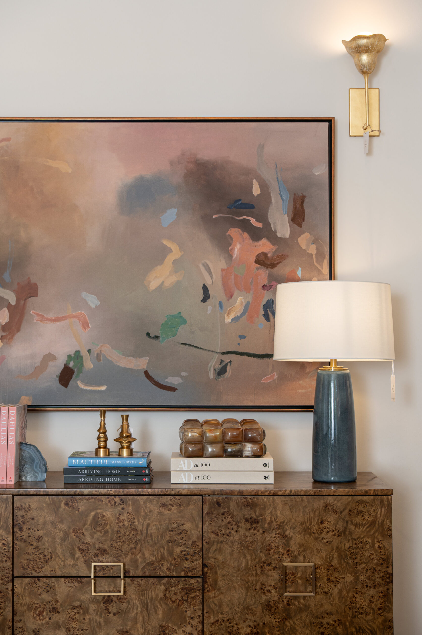 Modern abstract painting styled above a burl wood cabinet with a blue ceramic lamp, vintage brass candlesticks, and stacked coffee table books in a curated interior design vignette.