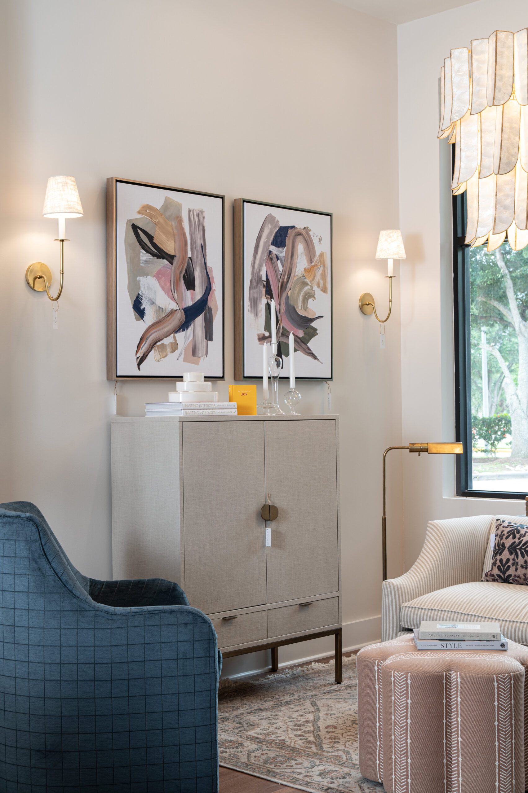 Modern living room corner with a cream-colored linen cabinet, two abstract art prints, brass wall sconces, and a mix of textured seating in blue and cream tones.

