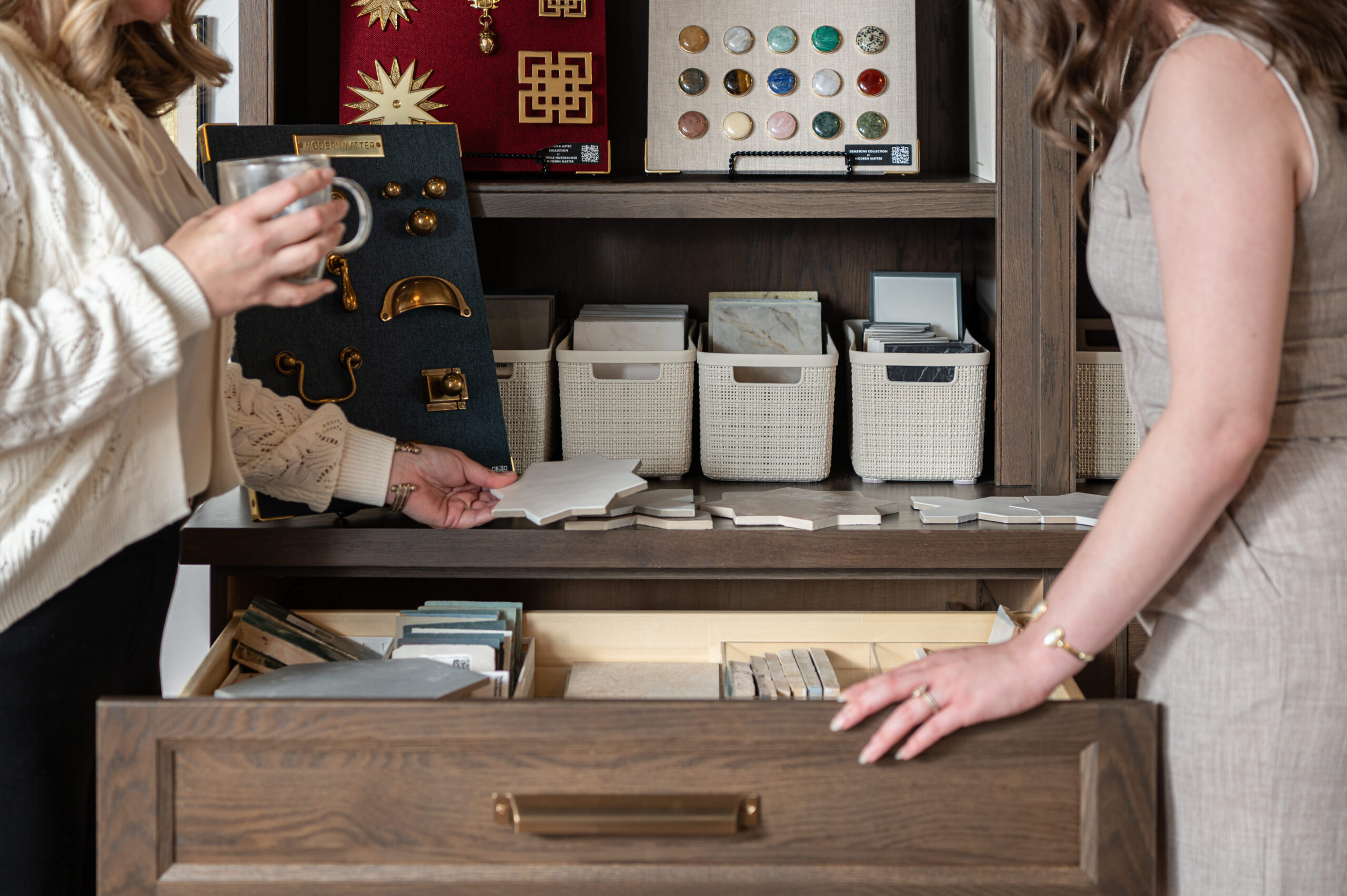 Hands of two designers reviewing tile samples in an interior design studio with brass hardware boards and marble options organized in woven bins.

