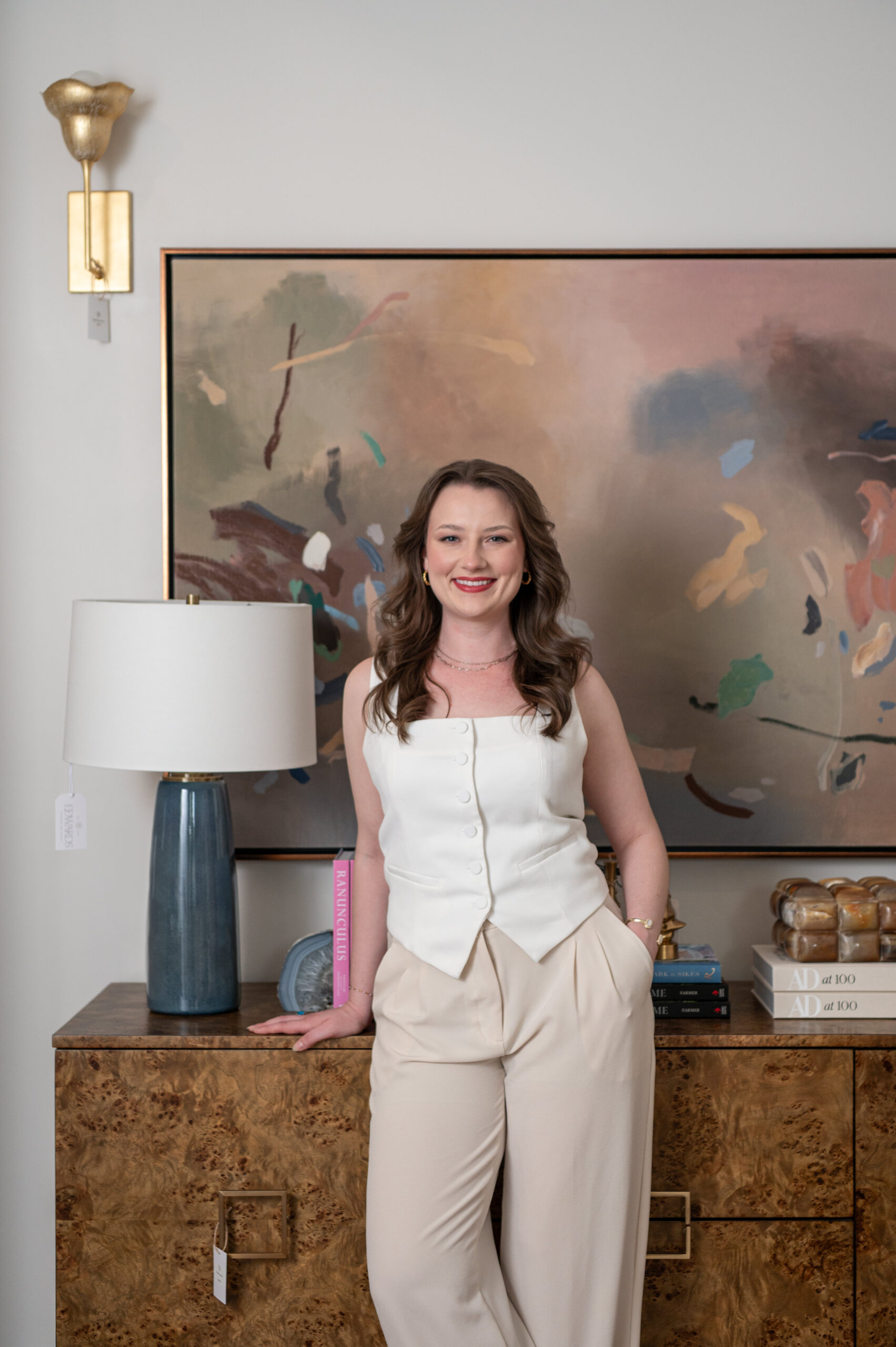 Smiling interior designer in a white top and cream trousers standing in front of a large abstract painting, styled console table with sculptural lamp and design books.

