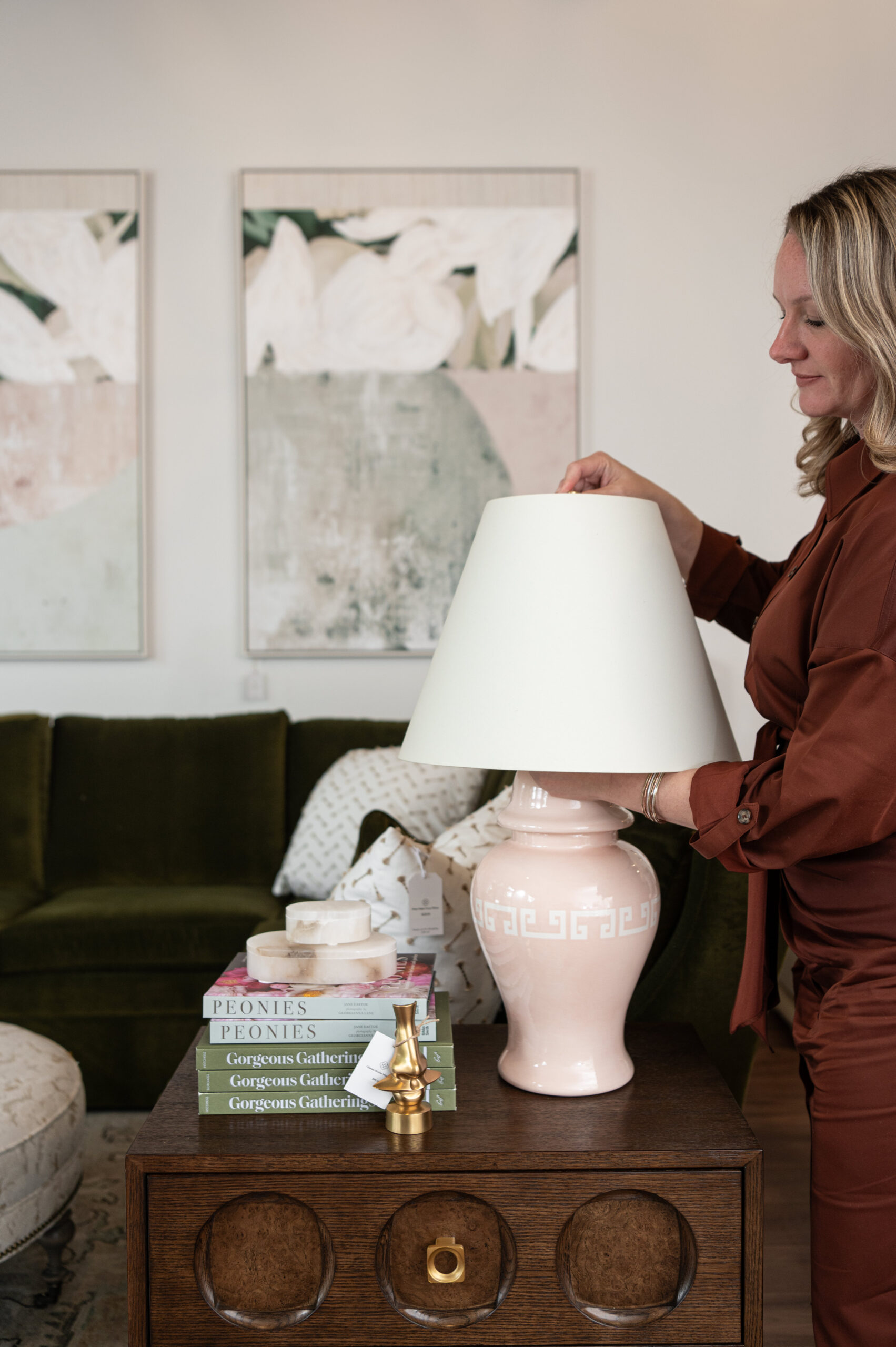 Interior designer adjusting a blush pink ceramic table lamp on a vintage wood side table, styled with coffee table books and a green velvet sofa in the background.

