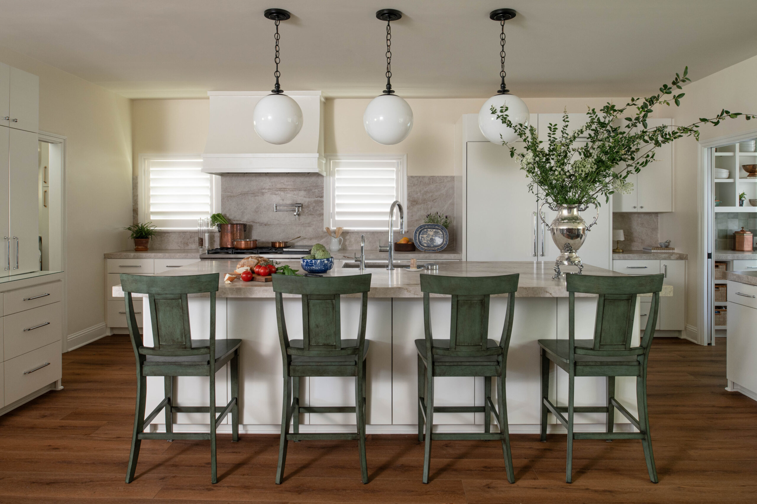 Wide kitchen view featuring green bar stools, globe pendant lights, and a dramatic floral arrangement in a silver urn.