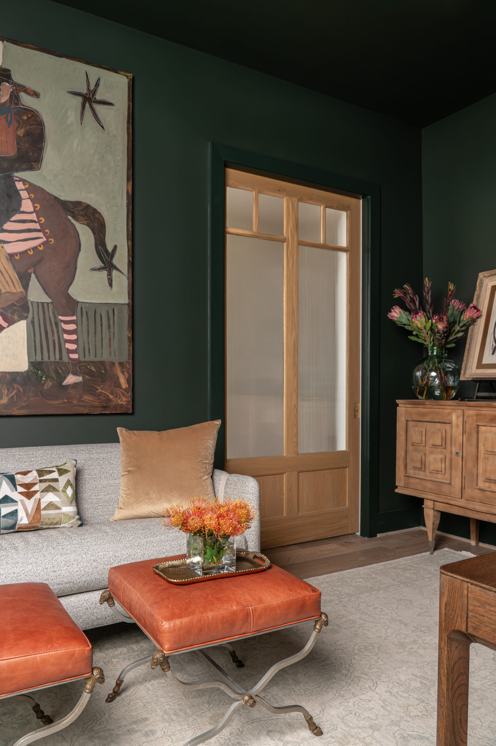 A living room featuring deep green walls, a wood-framed glass door, and a mix of leather, linen, and velvet fabrics that highlight the role of texture in interior design.