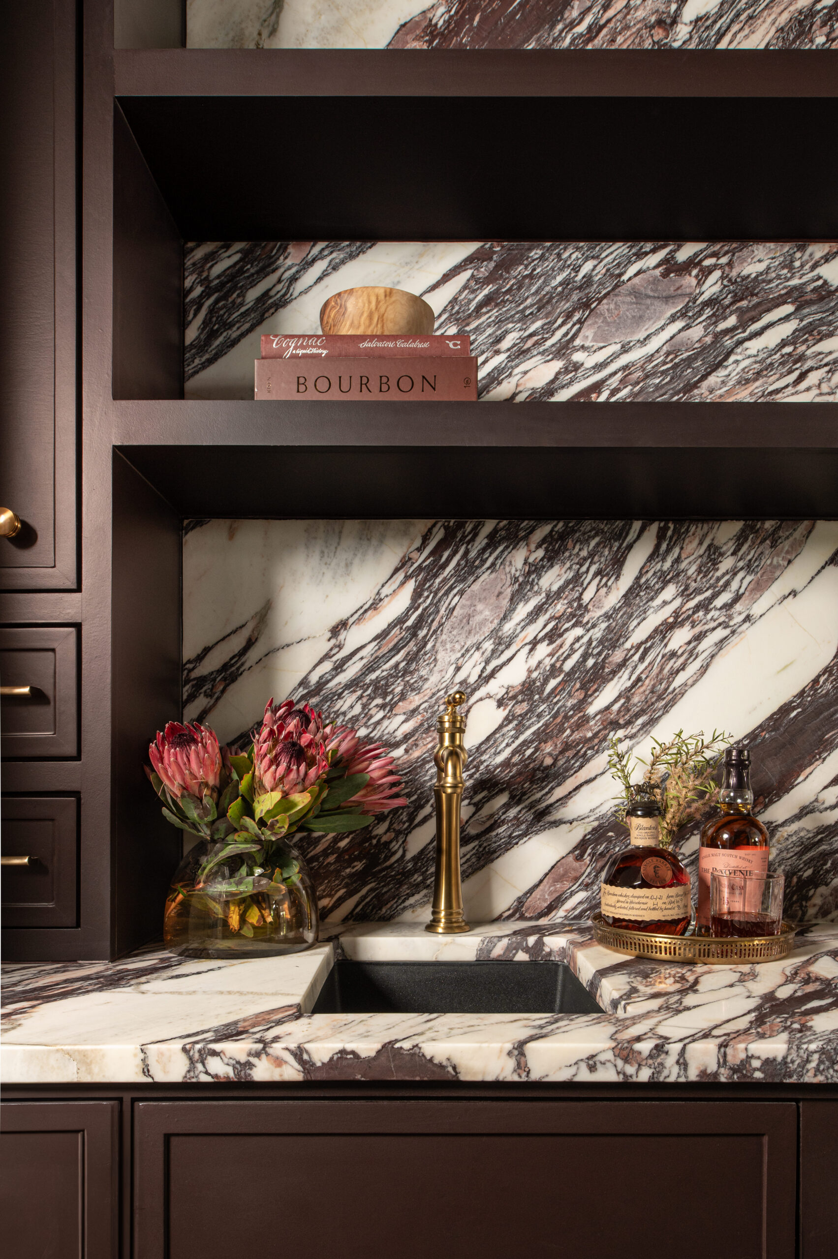 Marble backsplash and countertops add bold texture in interior design, paired with dark wood cabinetry, brass accents, and curated shelf styling.