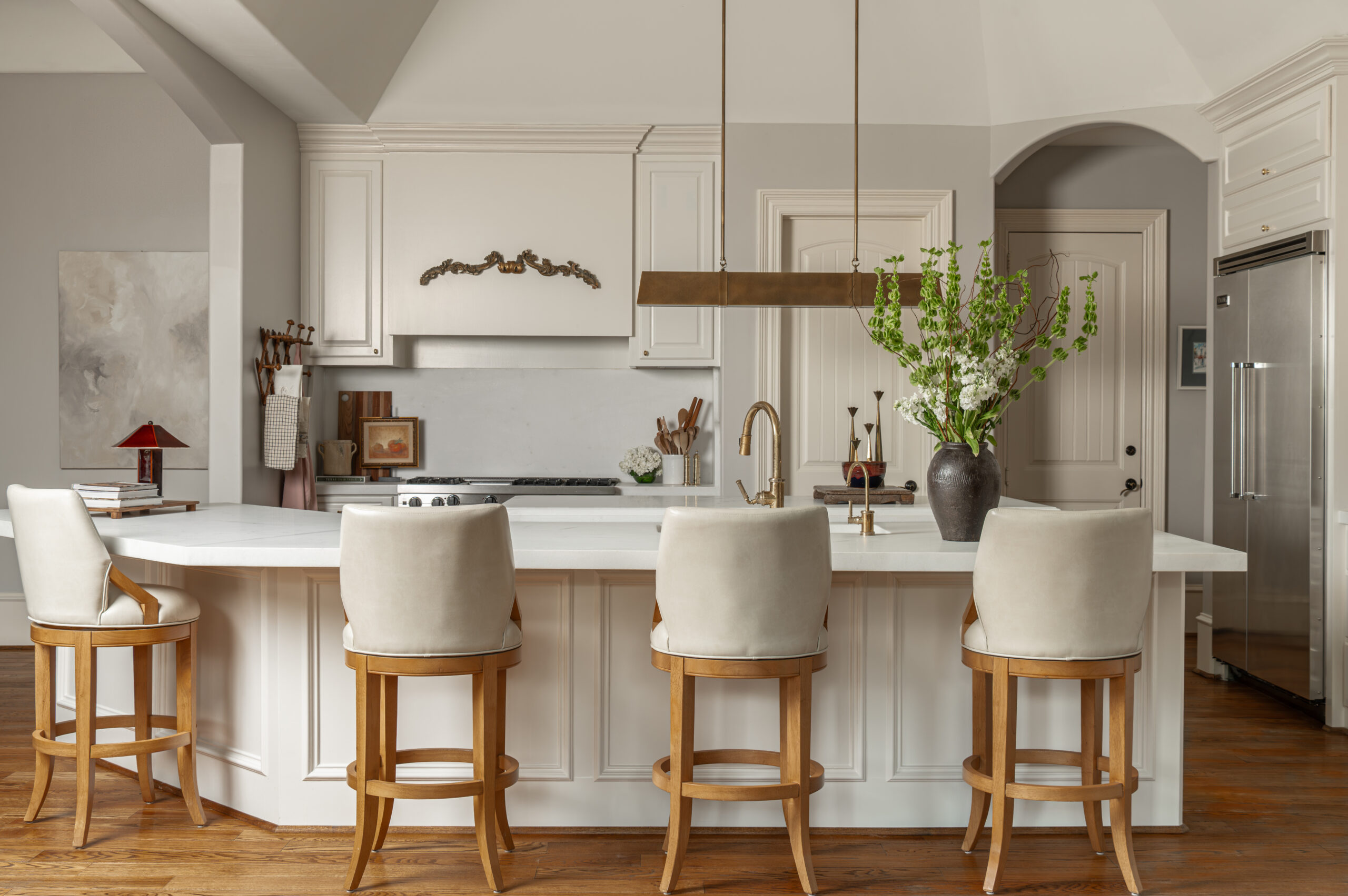 A wide view of the kitchen island and seating area, featuring a statement brass pendant light, white countertops, and four plush barstools—perfectly styled for a refined kitchen interior design shoot.