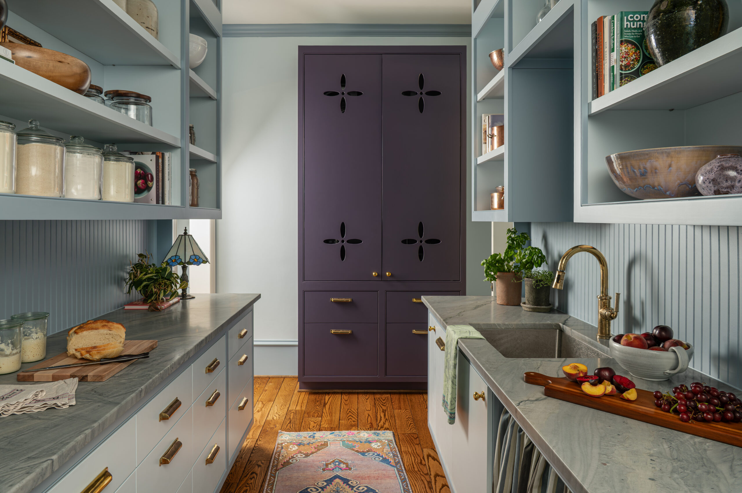 A moody and colorful prep kitchen with a deep plum pantry, open shelving filled with glass jars and cookbooks, and brass accents—designed to support everyday cooking in a high-functioning kitchen interior design.