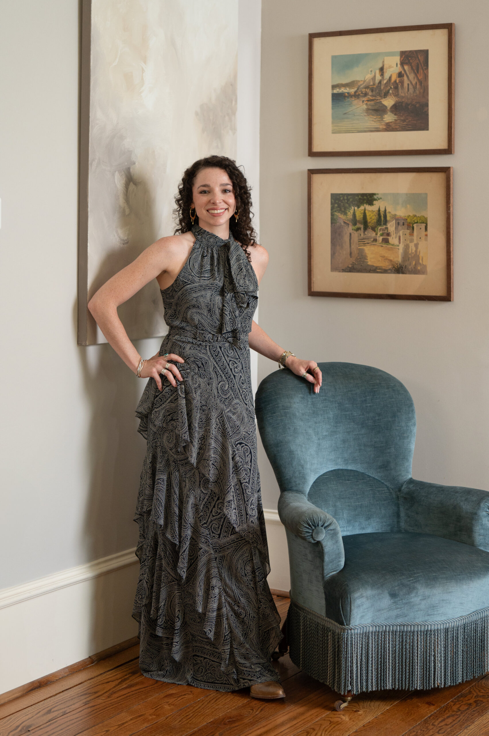 Melissa standing confidently next to a velvet armchair, with vintage wall art behind her—showcasing her timeless personal style and love for storytelling design.