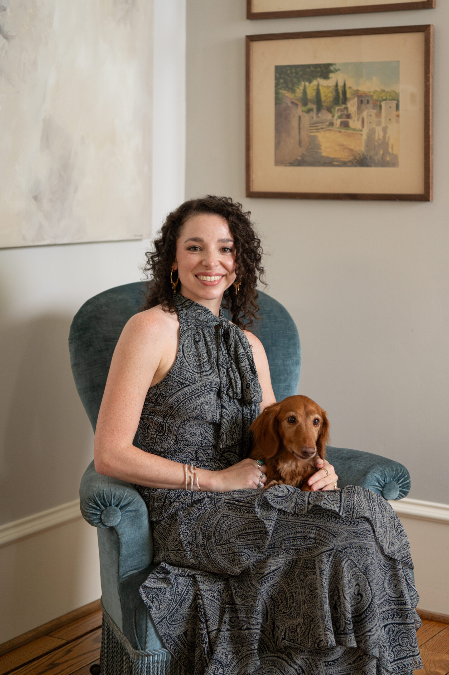 Melissa seated in a soft blue armchair holding her dog, radiating warmth and personality in the relaxed corner of the home.